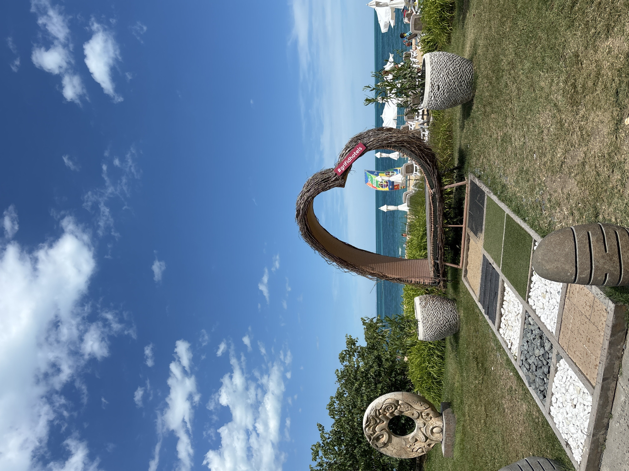 The image captures a sunny day at a beach resort in Varna, Bulgaria. The foreground features a decorative heart-shaped arch made of natural materials, likely a photo opportunity. Next to it, a display showcases different types of ground cover, including sand, gravel, artificial grass, and paving stones. Several large, decorative stone planters and sculptures add to the ambiance.
In the mid-ground, the beach is visible with rows of lounge chairs and umbrellas, suggesting a leisurely atmosphere. The blue sea stretches to the horizon, meeting a clear blue sky dotted with fluffy white clouds. In the distance, people can be seen relaxing on the beach, enjoying the seaside.
A small, red sign attached to the heart arch displays the hashtag "#grifidhotels". The overall scene evokes a sense of relaxation and holiday enjoyment on a bright, clear day.