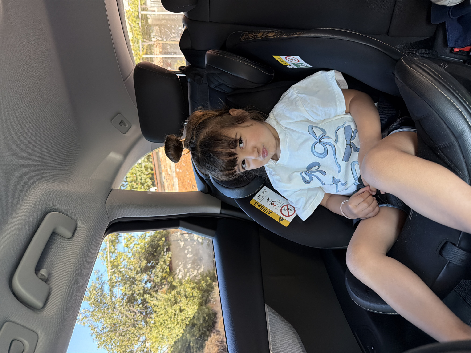 A young girl with her hair in a bun is sitting in a car seat in the back seat of a vehicle. She is wearing a white t-shirt with blue bows printed on it and denim shorts. The car seat has a yellow airbag warning sticker. The interior of the car is visible, including the headrests and a grab handle on the ceiling. Outside the car window, trees and a brick wall can be seen under a clear blue sky. The location is identified as Burgas, Bulgaria.