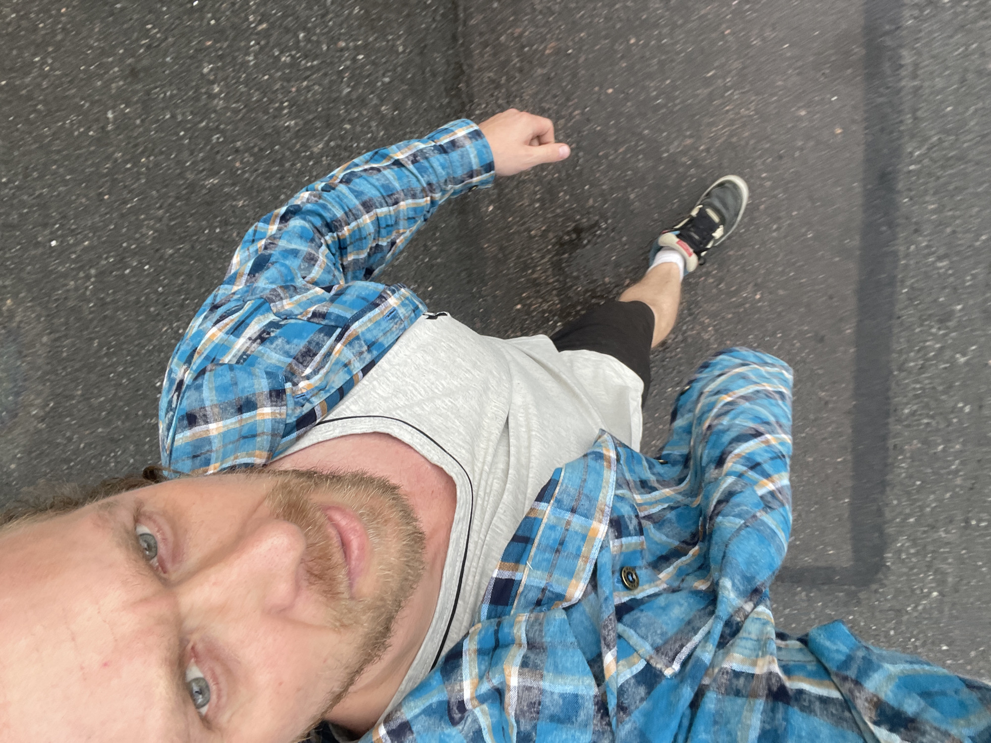 A man with light skin, blonde hair, and a beard is looking up at the camera. He is wearing a light gray t-shirt under an open blue plaid shirt. He is also wearing black shorts and dark sneakers with white socks. The man appears to be walking on an asphalt surface, possibly a street or sidewalk in an urban environment. The image is taken from a high angle, looking down at the man. The background is out of focus, but appears to be a gray asphalt surface with some scattered white specks, suggestive of gravel or debris. The lighting is overcast, creating soft shadows. The man's expression is serious, with his eyebrows furrowed. There are no other people or discernible landmarks in the image.
