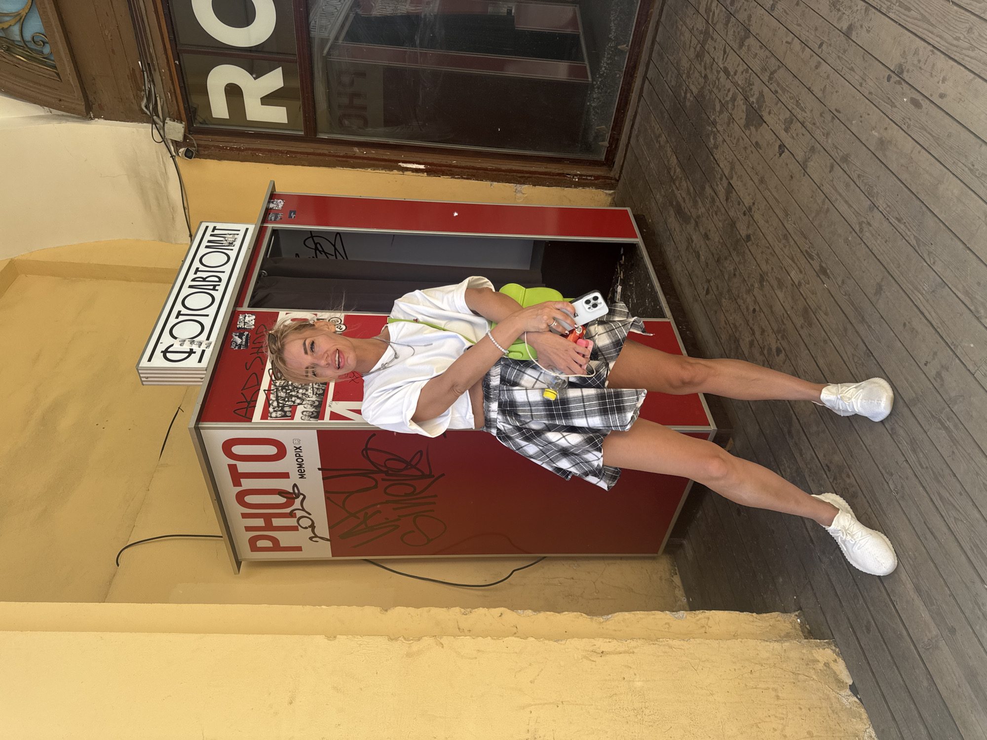 A woman is posing for a photo in front of a photo booth. She is wearing a white t-shirt, a black and white plaid skirt, and white sneakers. She is holding a smartphone and a bright green bag. The photo booth is red with white lettering that says "PHOTO" and "MEMORIES" in Russian. There is also graffiti on the side of the photo booth. The scene appears to be outdoors, with a wooden walkway and a yellow wall. The lighting suggests it is daytime, with the sun casting shadows. The woman has a smiling expression, appearing happy and playful. The setting is likely a public area, possibly a street or a courtyard in Saint Petersburg, Russia, given the context.