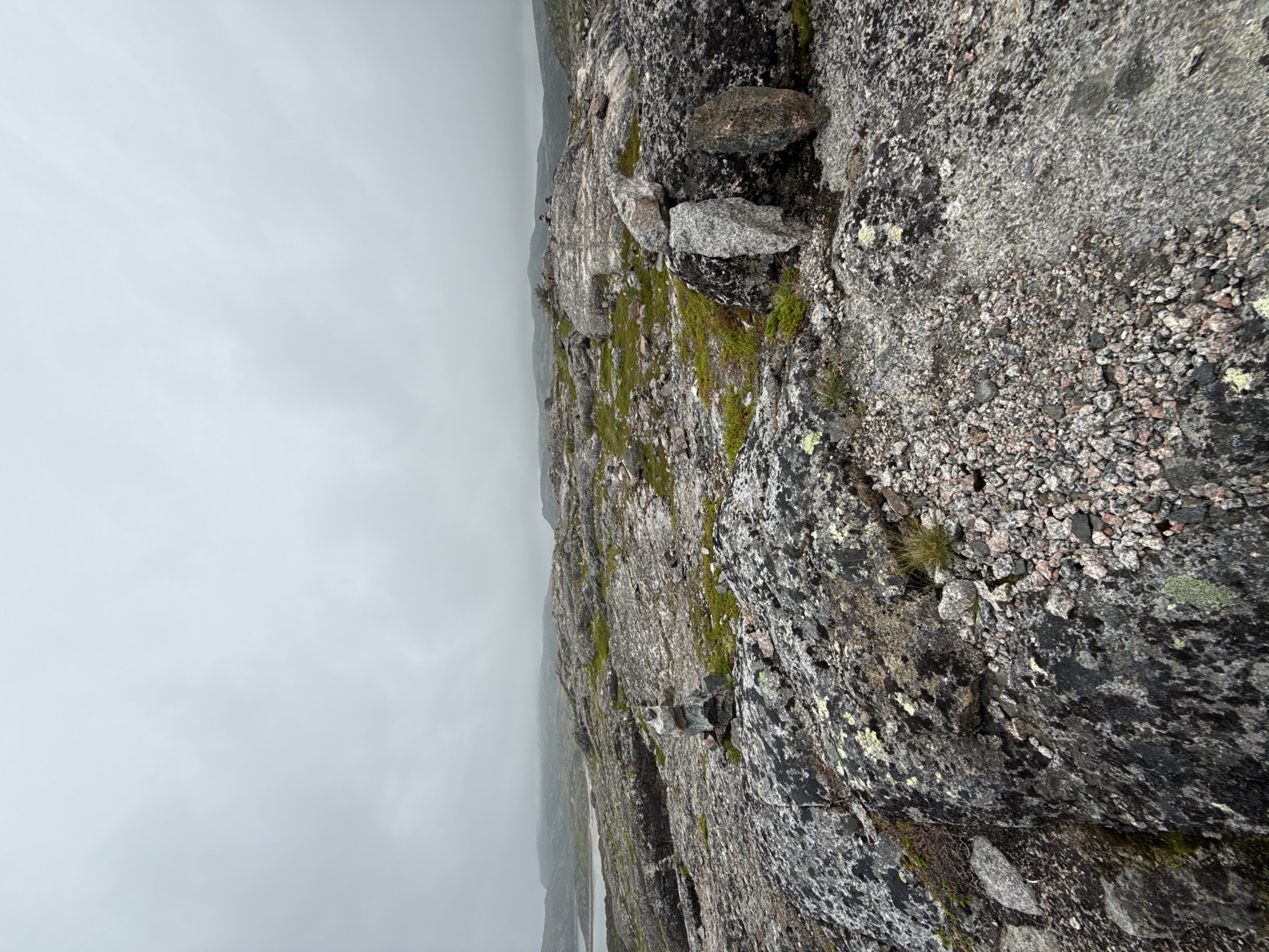 The image depicts a rocky, barren landscape under a cloudy sky, consistent with the environment found in Teriberka, Russia. In the foreground, large, dark gray rocks with lighter gray and pinkish speckles are prominent, covered in patches of green moss and lichen. Small, loose stones and gravel are scattered across the surface. A small cairn, a stack of stones, is visible to the left of the center, appearing to mark a path or viewpoint.
The middle ground transitions to a flatter, less rugged terrain interspersed with patches of green vegetation and scattered rocks. Further in the distance, a body of water, likely the Barents Sea, is visible, with a low, hilly coastline stretching across the horizon. The overall atmosphere is subdued, with muted colors and a lack of distinct shadows, indicating an overcast day. The scene is devoid of any people or animals, presenting a purely natural environment. The image captures a sense of remote, rugged wilderness typical of the Arctic coastal regions.