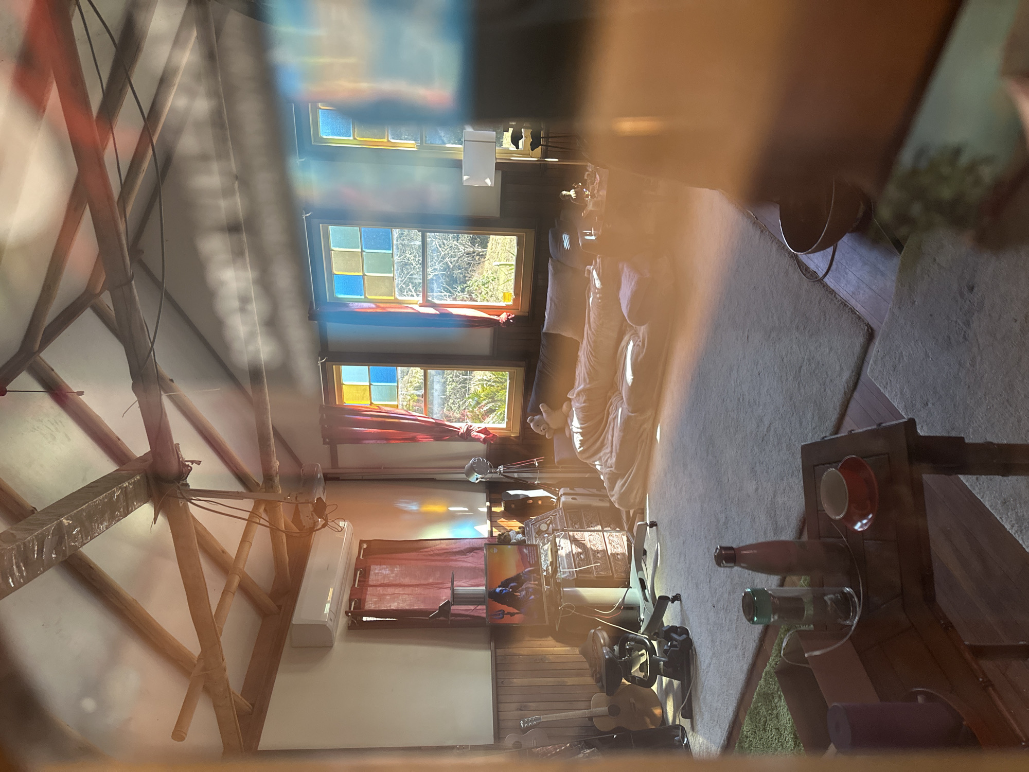 This image depicts the interior of a rustic, attic-like room, likely a living space or studio, bathed in warm, colorful sunlight streaming through stained-glass windows. The room features exposed wooden beams on the vaulted ceiling, an air conditioning unit mounted high on a wall, and dark wooden wainscoting.
In the foreground, a wooden table holds a red teacup and saucer, a water bottle with a green lid, and a dark-colored thermos. Adjacent to this, a purple yoga mat stands upright, and a brown cardboard box is visible. A large, plush rug covers a significant portion of the floor.
To the left, several acoustic guitars are propped against the wall. A desk with a computer monitor displaying an image of a serene figure, likely a deity, is situated behind a massage chair.
The central area of the room is dominated by a large, dark-colored couch or daybed, covered with blankets and pillows. A person, partially obscured by blankets, appears to be lying on the couch, their body relaxed.
In the background, through the stained-glass windows, a sunlit outdoor scene with trees and vegetation is visible. A floor lamp with a white rectangular shade stands near the couch. The overall ambiance is peaceful and bohemian, with the light casting vibrant, prismatic patterns across the room. The location in Thora, Australia, suggests a potentially coastal or rural setting, contributing to the natural light and serene atmosphere.