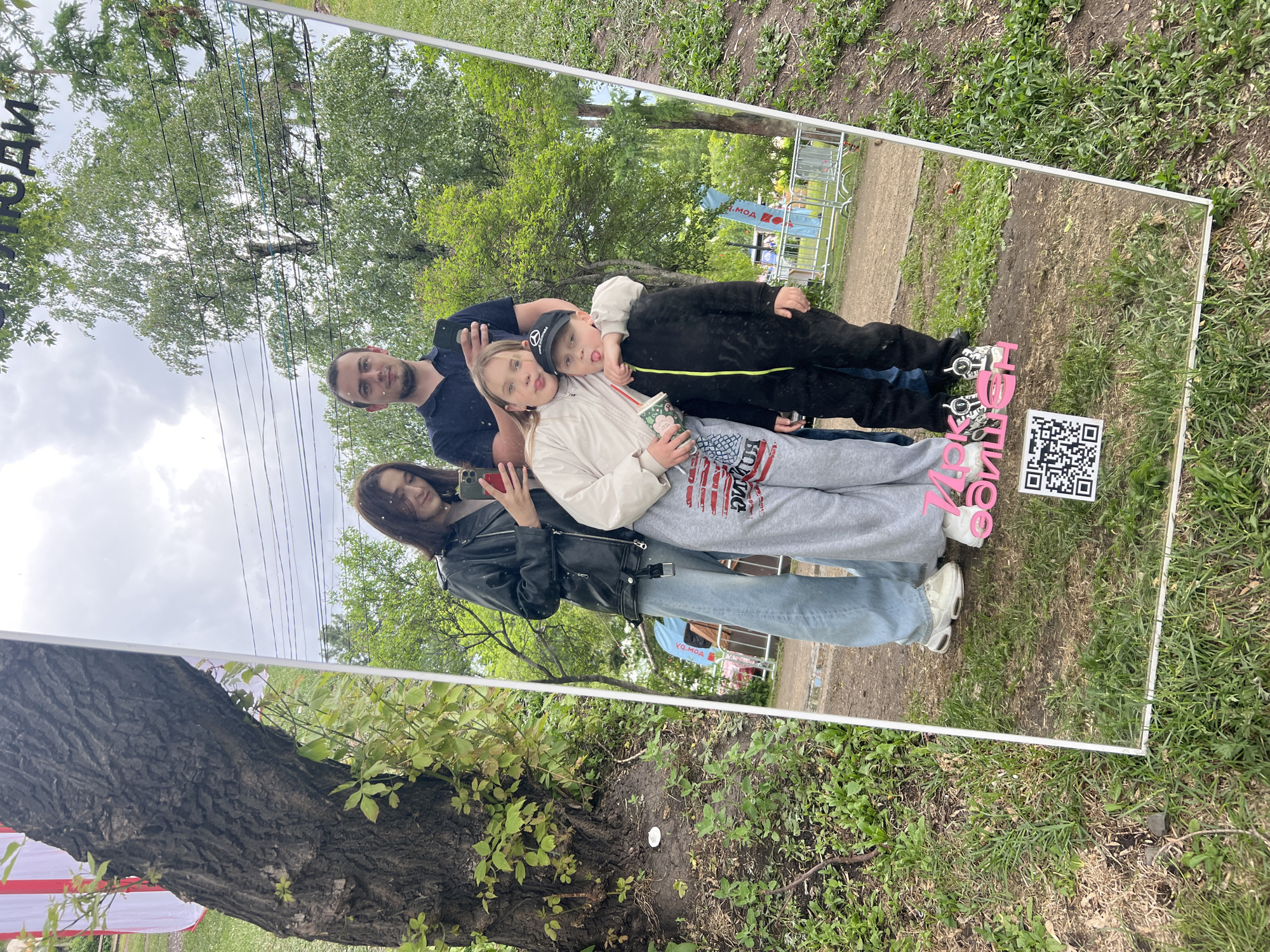 This outdoor photograph depicts a family of four reflected in a large, vertically oriented mirror with a white frame, situated on grassy ground in Irkutsk, Russia.
The mirror's reflection showcases an adult male, an adult female, a pre-teen female, and a young male child. The adult female, positioned on the left, wears a black leather jacket, a light top, light blue wide-leg jeans, and white sneakers, holding a dark smartphone with a red case. The adult male, with a beard, wears a dark polo shirt and dark trousers, holding a dark smartphone, with his right arm around the pre-teen female. The pre-teen female, centrally located, wears a light-colored long-sleeved top, grey sweatpants with a red, white, and blue graphic, and white sneakers. She holds a patterned cup and sticks her tongue out. The young male child, on the right, wears a black baseball cap with a white Mercedes-Benz star logo, a black full-body tracksuit with neon yellow zipper trim, and dark sneakers. His left arm is around the pre-teen female's shoulder, and he is also sticking his tongue out.
Reflected in the top portion of the mirror is the Russian word "люди" (people). In the lower portion, pink block letters spell "14К ЗАКУПЩИК" (14K BUYER/PURCHASER), alongside a black and white QR code.
The environment outside the mirror includes a large tree trunk with textured bark and green foliage on the left. A red and white striped fabric structure is partially visible behind the tree. The reflected background beyond the family shows a cloudy sky, green trees, overhead power lines, a dirt path, white metal barriers, and a blue banner with white Cyrillic text (partially reading "МОА") on the right. The ground in the immediate foreground and surrounding the mirror is covered with green grass and patches of soil.