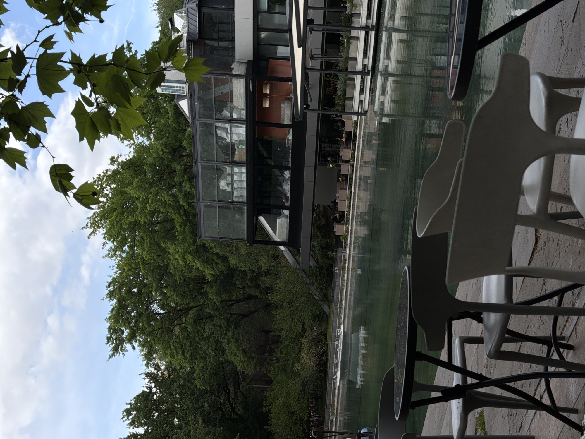The image depicts an outdoor cafe scene in Yerevan, Armenia. The cafe is located next to a body of water, possibly a canal or decorative pool, which reflects the building's structure.
The building appears to be a modern architectural design featuring glass windows and steel beams. Green trees and foliage are visible around the building, suggesting a park-like setting.
Several white plastic chairs are arranged around tables. The tables have black frames and glass tops. The scene is deserted. The sky is partially visible, with a mix of clouds and blue.