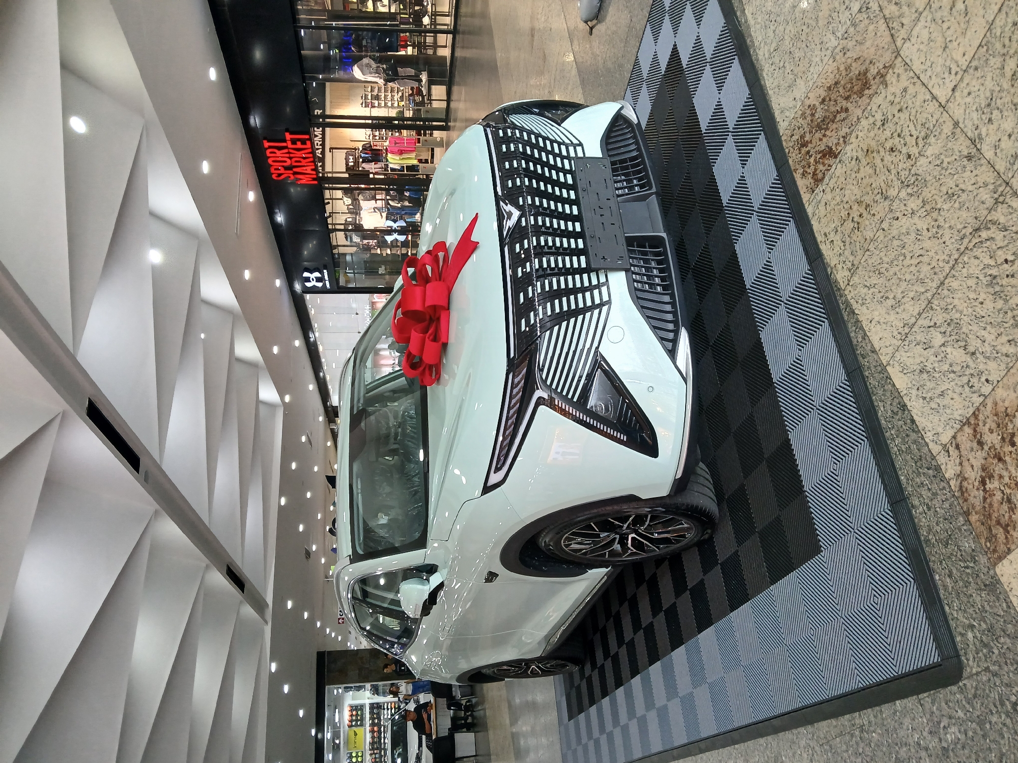 A white vehicle with a large red bow on its hood is parked on a checkered black and gray platform inside a commercial space. The car is positioned in front of a "Sport Market" store in the city of Ciudad Guayana, Venezuela. The location is a shopping mall, evident from the store displays and ceiling lights. Two people are faintly visible near the storefront, suggesting activity within the mall. The floor transitions from the checkered platform to a light-colored marble or tile surface.