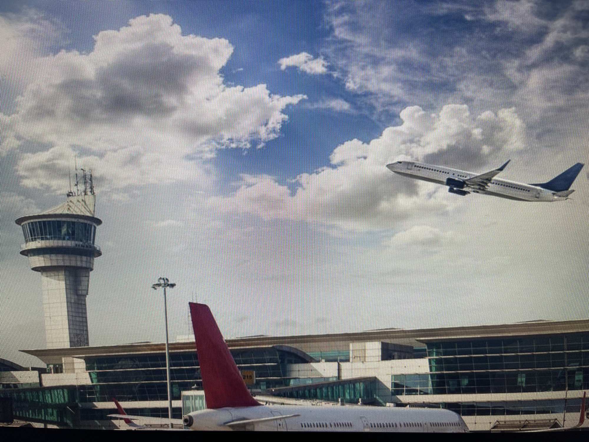 The image captures an aerial view of Varna Airport in Varna, Bulgaria. A modern airport terminal with large glass windows is visible in the foreground, with the tail of a red-tailed aircraft positioned prominently. In the background, a control tower stands tall, and another airplane is seen ascending into a sky filled with white, fluffy clouds against a blue expanse. The scene depicts a busy airport environment under a clear sky.