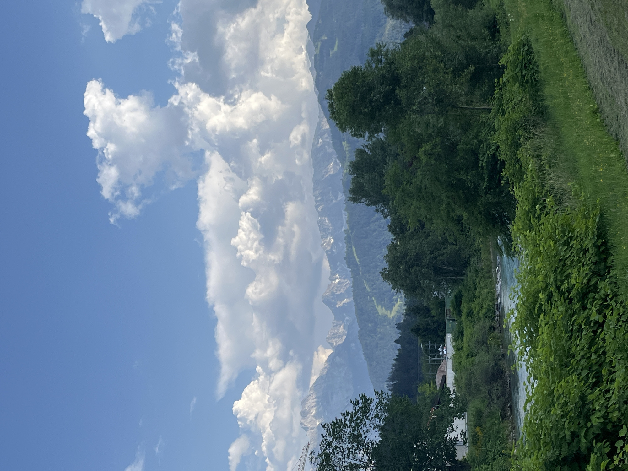 The image captures a picturesque landscape with a clear blue sky dotted with fluffy white clouds. In the distance, a majestic mountain range rises, its peaks partially covered in snow, suggesting a high altitude. Verdant green forests cover the lower slopes of the mountains, and a winding ski slope is visible on one of them, implying a location in a mountainous region known for winter sports.
In the foreground, a tranquil river flows, its water a refreshing turquoise hue. Lush green foliage lines the riverbanks, with dense trees and bushes creating a vibrant natural border. A few buildings are partially visible among the trees on the left side of the image, indicating human habitation in the vicinity. The overall scene evokes a sense of natural beauty and serenity, characteristic of a rural or semi-rural setting within a mountainous area. Given the context of Garmisch-Partenkirchen, Germany, this scene is likely situated in or around this Bavarian town, known for its stunning alpine scenery and outdoor recreational opportunities. The presence of the ski slope specifically points to the proximity of ski resorts common in this region.