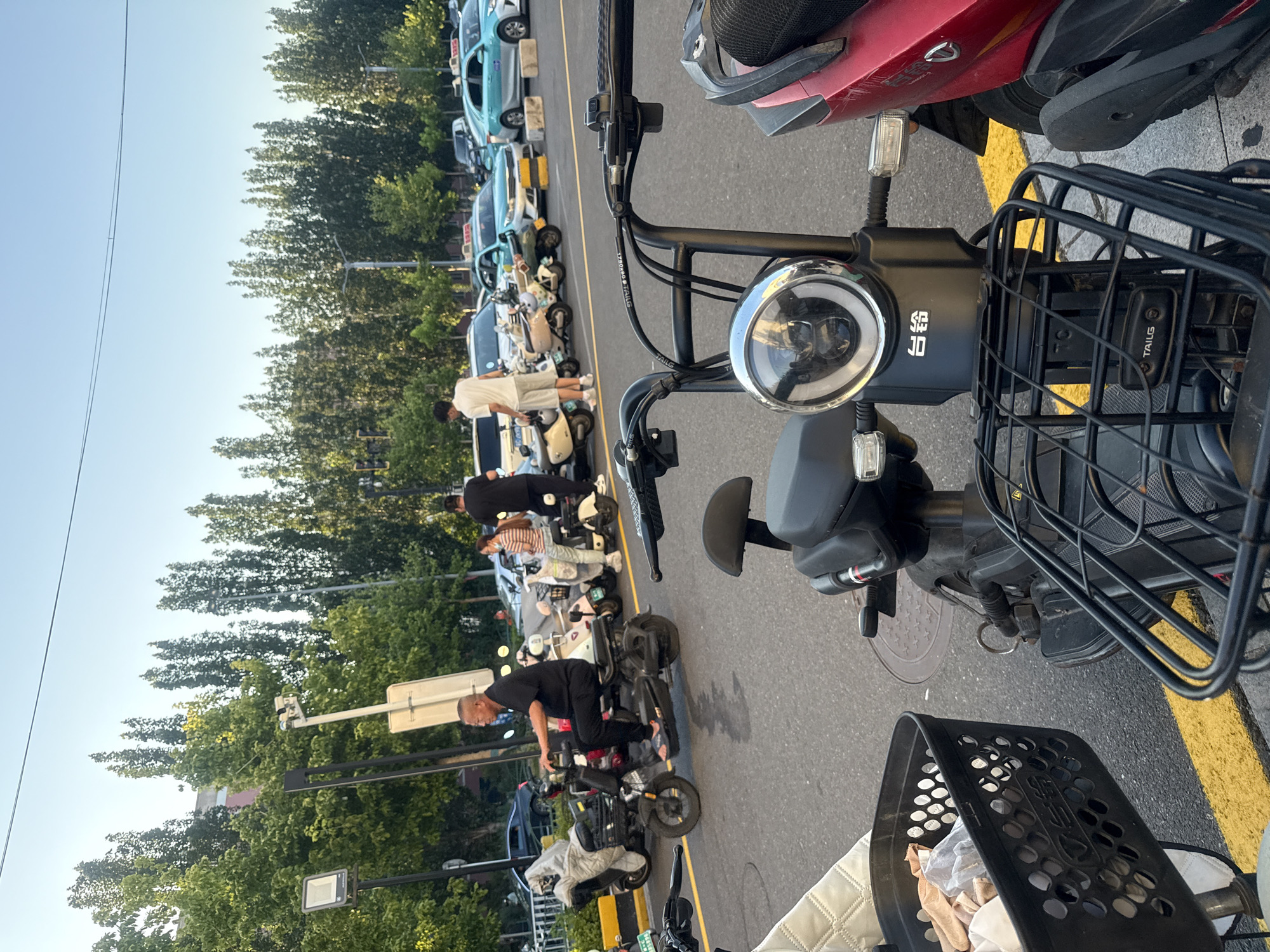 The image captures a street scene in Qinhuangdao, China, likely during daylight hours with a clear sky. In the foreground, two electric scooters are parked, one black and one red. The black scooter has a wire basket filled with items and a visible logo on its front. The red scooter is partially visible, showing its side and rear.
The midground reveals several people and a variety of parked electric scooters and motorcycles. An older man in a black shirt and pants is riding a black electric scooter. A woman in a striped shirt and jeans is standing next to a beige scooter. Another person in a white shirt and light shorts is standing near a white car.
In the background, a row of tall, slender trees lines the street, with some buildings visible behind them. Several cars, including teal-colored taxis, are parked or moving in the distance. A traffic light is also visible on a pole. The overall impression is of a busy urban environment with prevalent personal electric transportation.