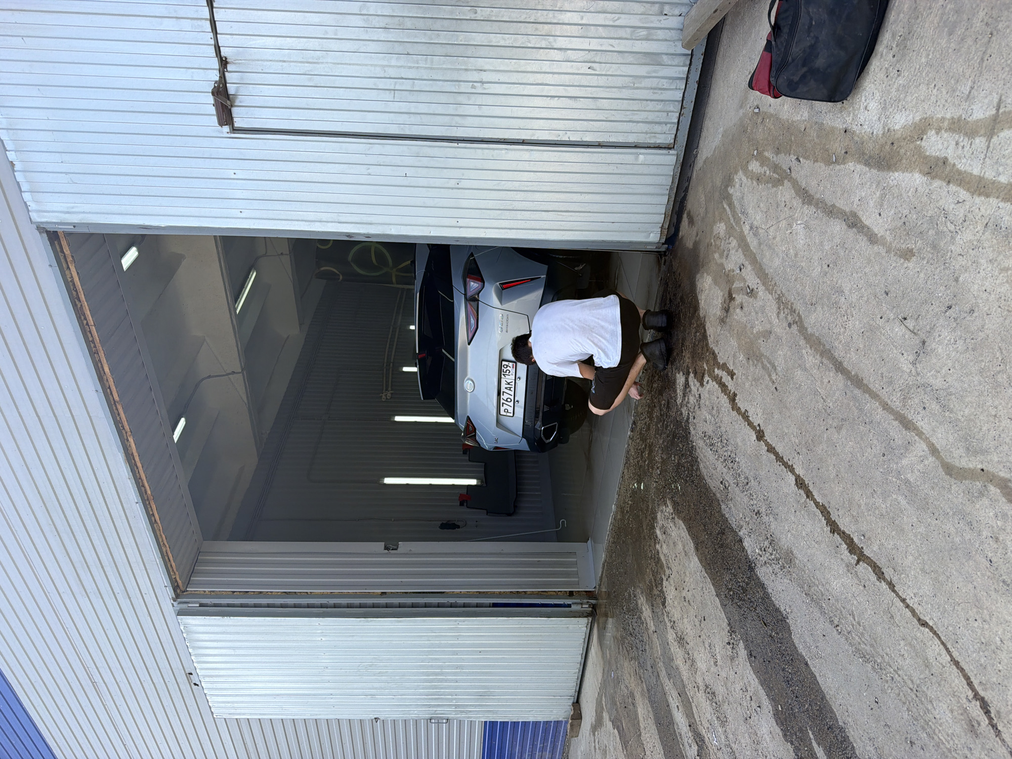 In the city of Chaikovsky, Russia, a person wearing a white t-shirt and dark shorts is crouching on the wet, concrete ground in front of an open garage-like structure. Inside the structure, a light-colored car, possibly a crossover SUV, is parked. The car has a Russian license plate that reads "P767AK 159". The garage is illuminated by fluorescent lights. The ground outside the garage is covered with water puddles and tire tracks. To the right of the image, a black and red duffel bag is placed on the ground. The overall scene depicts a car being washed or maintained in an open garage setting.