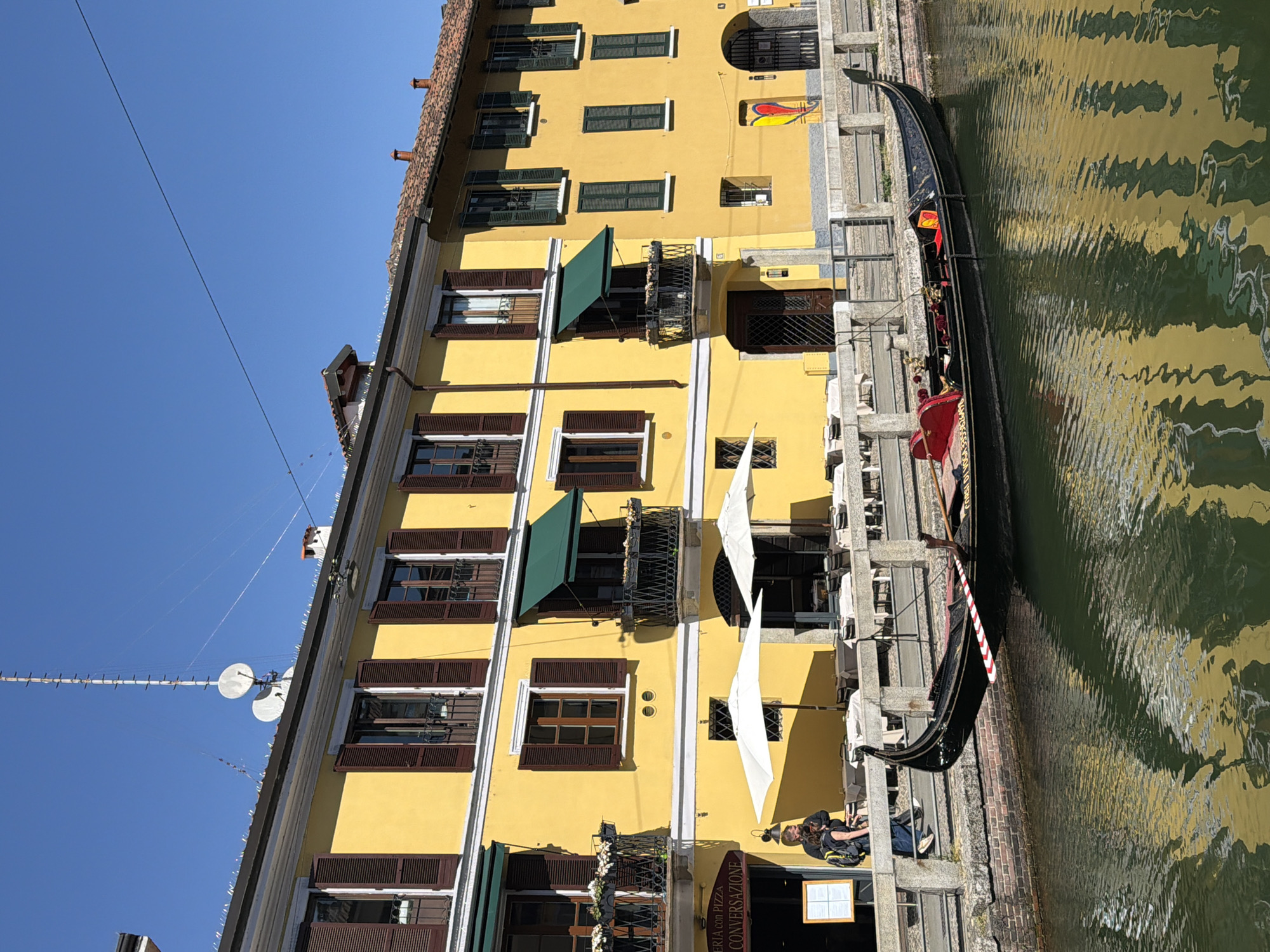 The image shows a canal scene in Milan, Italy. A gondola is moored alongside a building with a yellow facade. The building has multiple stories, with windows featuring brown shutters, some with green awnings, and small balconies. A tall antenna with satellite dishes is attached to the building. Tables and chairs are set up along the canal side, and a person is visible near a menu board for a restaurant. The water in the canal is green and reflective. The sky is clear and blue.