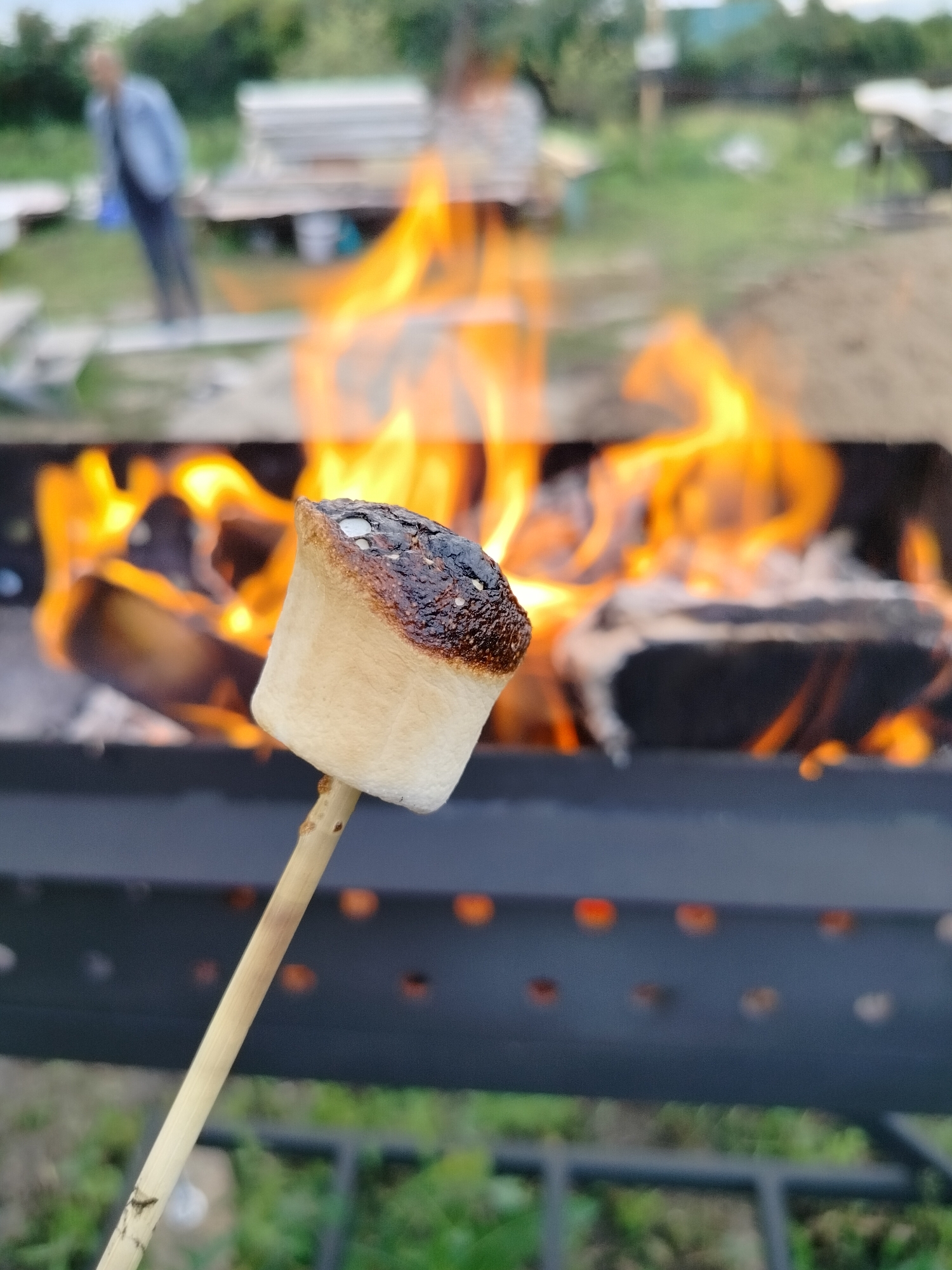 This media file captures a close-up of a marshmallow being toasted over an open fire. The marshmallow, impaled on a wooden skewer, is prominently featured in the foreground, with its top half nicely browned and slightly charred, indicating it's been held near the flames. In the background, a fire is burning brightly within a dark metal grill or fire pit. The flames are a vibrant orange and yellow, illuminating the scene.
Beyond the fire pit, the setting appears to be an outdoor, rural area. Blurry figures of people and structures can be discerned. One person, wearing a light blue jacket and dark pants, is visible in the upper left quadrant, standing at a distance. There are also indistinct shapes that suggest wooden structures or benches. The ground is covered with green grass and what appears to be sand or dirt. The overall impression is that of a casual outdoor gathering or a moment of relaxation. The lighting suggests it could be daytime, possibly late afternoon or early evening, given the warmth of the fire and the ambient light. The location context provided is Вазерки, Russia, which aligns with the rustic, outdoor atmosphere. There is no visible text in the media file.