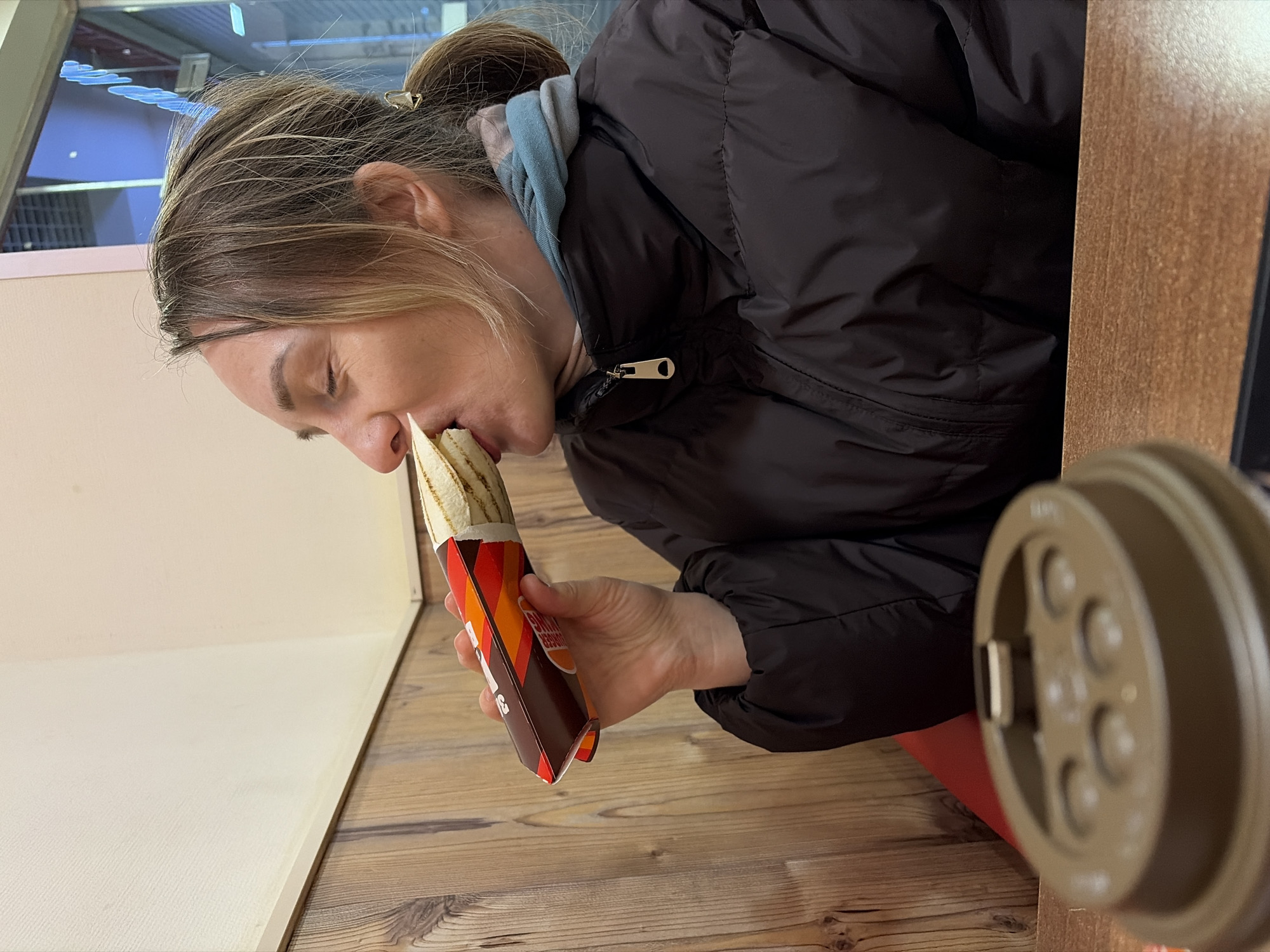 A woman with fair hair pulled back in a bun, wearing a black puffy jacket and a blue hooded sweatshirt, is seated at a wooden table and eating a wrap from Burger King. The restaurant appears to be located in Izhevsk, Russia.