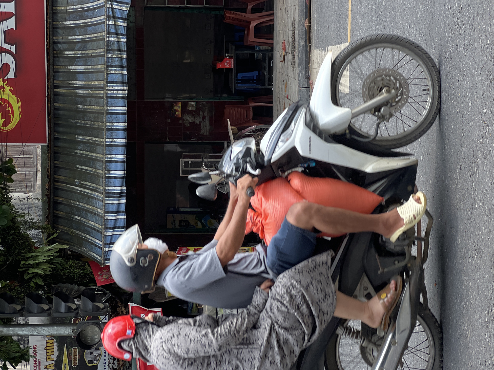 The image depicts a street scene in Tân Quang Commune, Vietnam, with two individuals riding on a white Honda scooter. The person in the front, driving the scooter, wears a grey polo shirt, blue shorts, a dark helmet with a clear visor, and a white face mask. They are carrying two large, orange, sack-like items strapped behind them. The passenger in the back is wearing a long-sleeved grey patterned garment, a red helmet, and a black face covering that extends up to their hairline. The scooter is traveling on an asphalt road, with a white pedestrian crossing line visible.
In the background, a building with a blue and white striped awning covers a storefront. The storefront features dark windows and red tiled walls. A sign with red and yellow lettering is visible above the awning, appearing to be advertising a product. Traffic lights are mounted on a pole to the left of the image, with multiple black signal heads. The environment suggests an urban or semi-urban setting. The overall lighting indicates it is daytime.