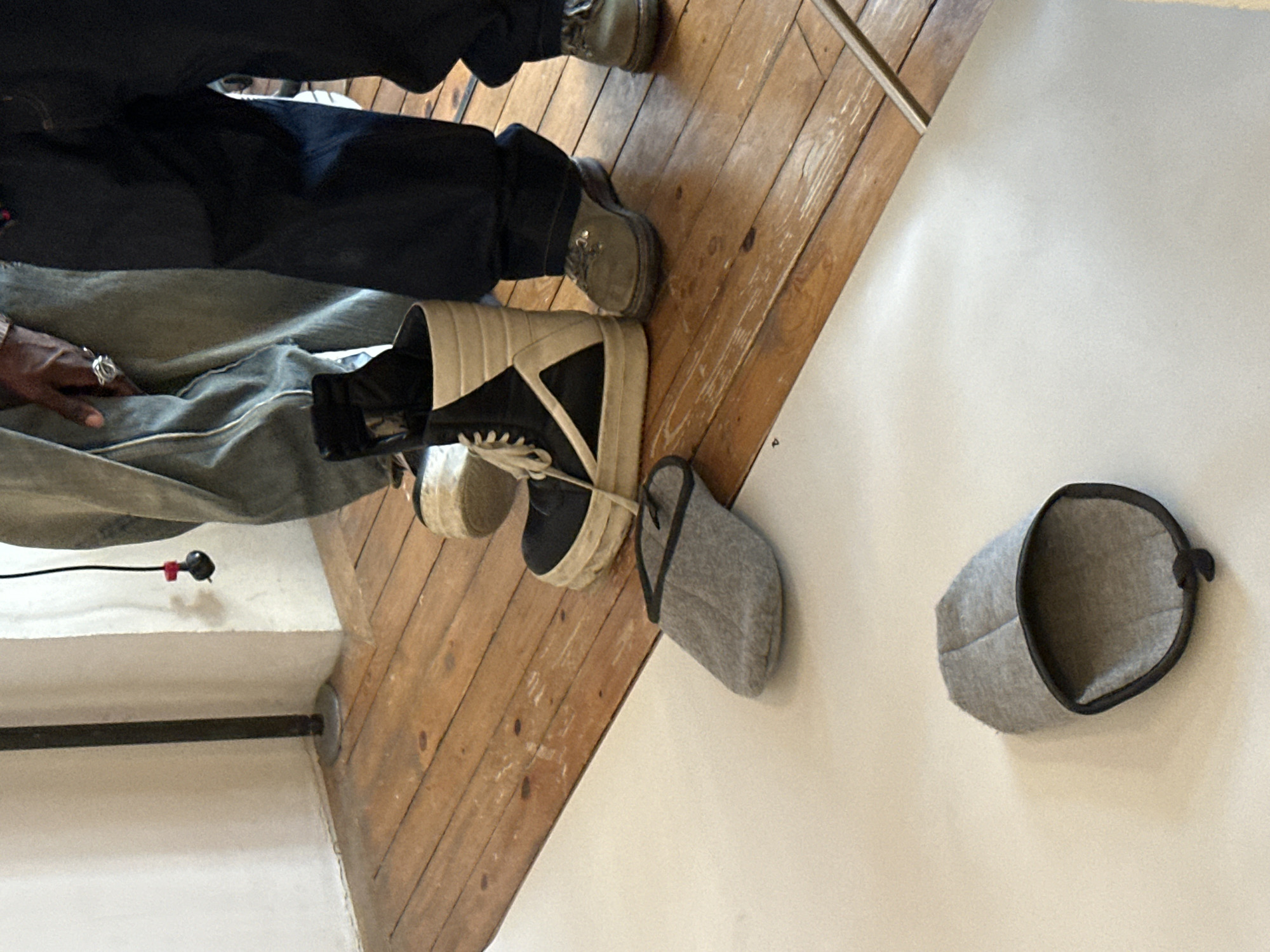 The image captures a scene indoors, showcasing a wooden floor transitioning to a white surface, likely a wall. Two pairs of feet are visible, one clad in casual shoes and denim jeans, and another wearing high-top sneakers. A light gray fabric object with a dark trim, cylindrical in shape, and seemingly open at both ends, rests on the white surface. Another, similar object is partially visible beside the sneakers on the wooden floor. A hand with a ring is noticeable resting on denim fabric. The location cannot be determined based on the visual information provided.