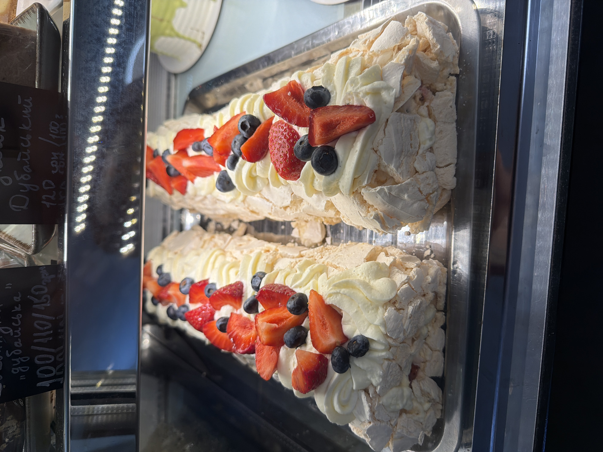 The image shows two dessert rolls displayed in a metal tray, likely in a bakery or cafe display case. The rolls are made of meringue, filled with cream, and topped with fresh strawberries and blueberries. The meringues have a textured, airy appearance, with visible cracks and crisp edges. The cream is piped in decorative swirls, and the fruit is arranged generously on top.
One of the rolls is more in focus and fills the majority of the frame. The second roll is visible behind it, slightly out of focus, suggesting they are presented side-by-side. The lighting suggests an indoor setting, possibly artificial light from the display case, as indicated by the bright highlights and reflections on the metal surfaces. The overall impression is one of fresh, appealing desserts.
There is visible text on a black tag in the lower left of the image. The text is handwritten in white chalk. It appears to be a product name and price. The text is partially obscured but includes "Дубайська" and "100/110/150". The orientation of the text suggests the tag is meant to be read from a different angle.