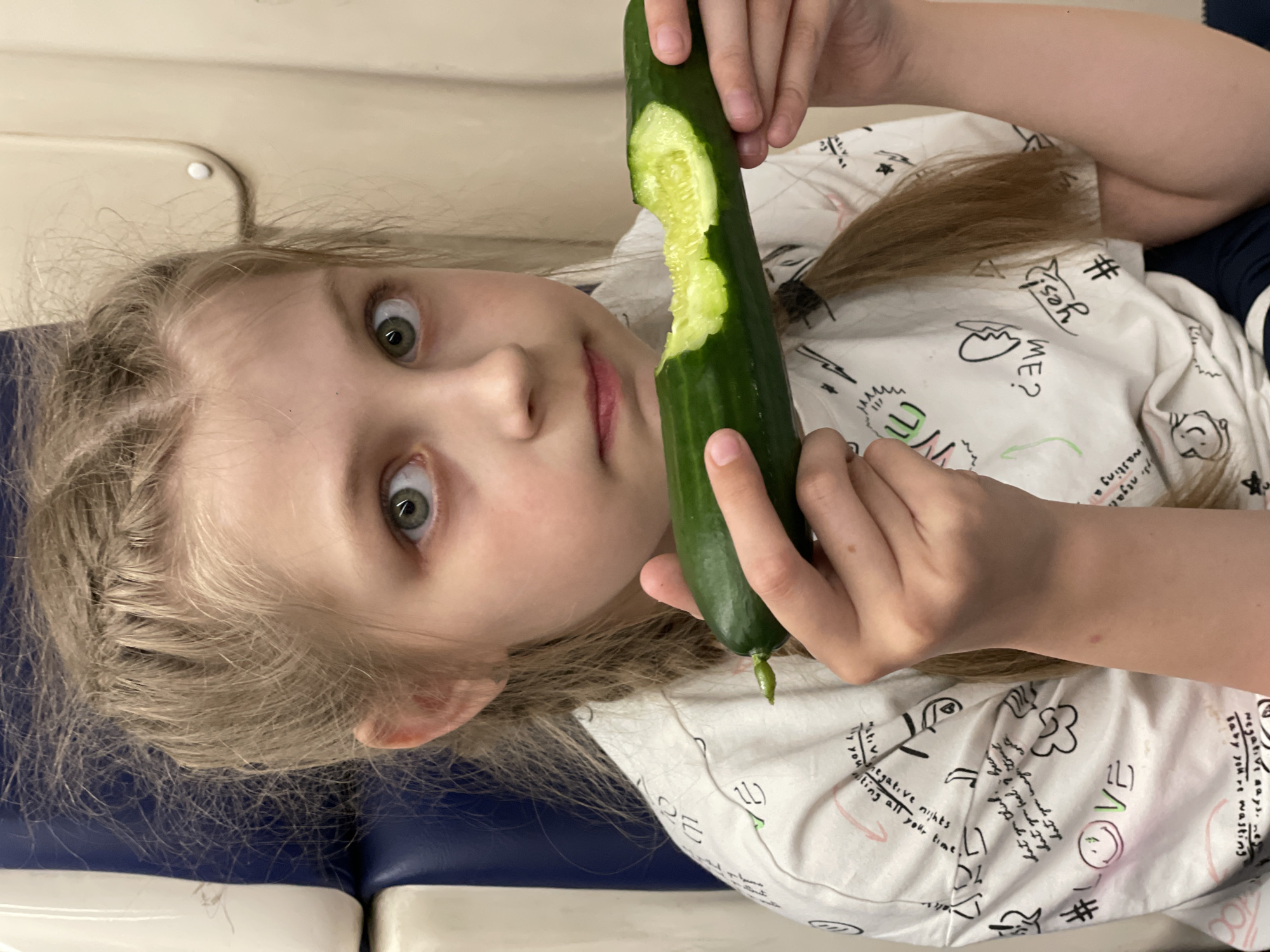 A young girl is lying down, looking at the camera and holding a partially eaten cucumber. She has blonde hair and is wearing a white shirt with doodles. She is indoors, on a surface that appears to be cushioned with blue and white sections. There is no evidence in the image to determine that the image was taken in Agryz, Russia.