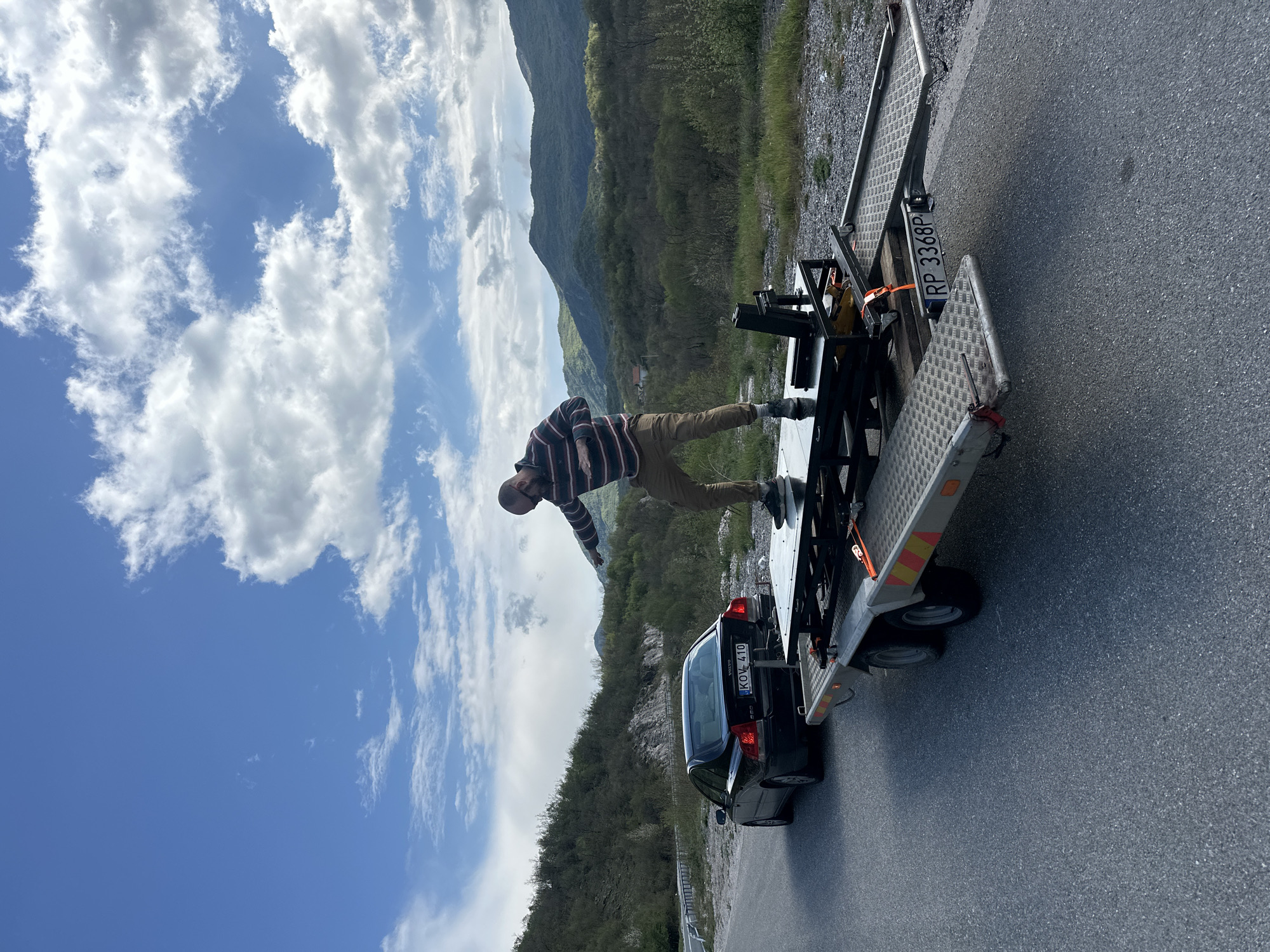 The image shows a man standing on a car trailer parked on a road in Očinići, Montenegro. The man is wearing a striped shirt and khaki pants. He is balancing with his arms outstretched. The trailer has a black car secured on it. The trailer's license plate reads "RP 3368P". The car's license plate reads "KOV 410". The background includes mountains, trees, and a cloudy sky.