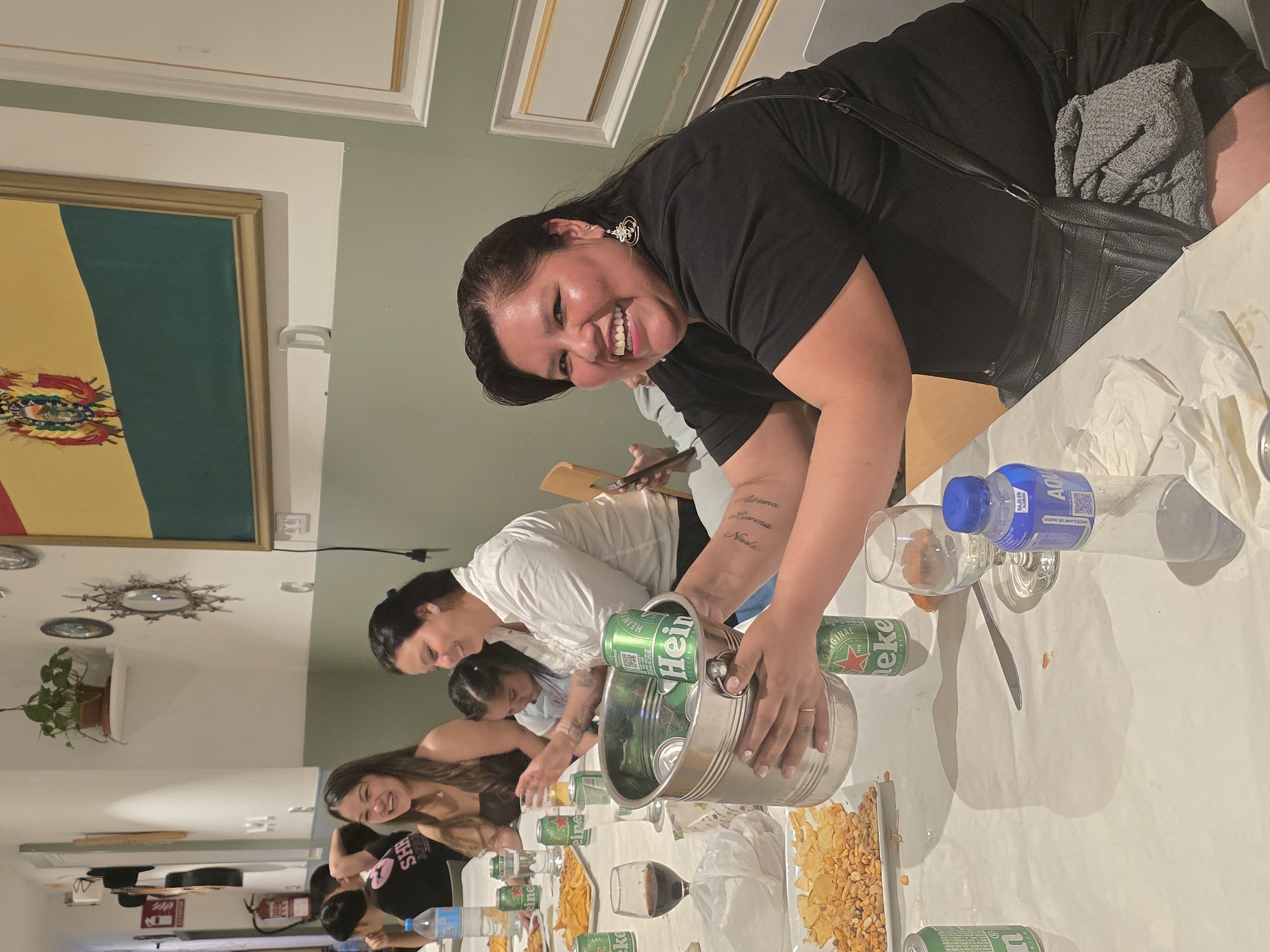 An indoor social gathering, likely an evening party or casual meal, is captured from a slightly elevated perspective in L'hospitalet De Llobregat, Spain. The main subject, a woman with dark hair wearing a black shirt and large circular earrings, smiles broadly at the camera as she leans over a long table. Her arm displays tattoos with names like "Alexa," "Lorena," and "Nicole."
The table, covered with a white cloth, is laden with food and drinks. Snacks include potato chips and peanuts in white dishes. Numerous green Heineken beer cans, some in a silver ice bucket, are visible, along with wine glasses, and blue-capped water bottles labeled "AQU," some indicating "BAJO EN SODIO" and "RECIÉNLAME DE NUEVO." Another woman in a white shirt is seen holding the ice bucket, while other individuals, mostly women, are seated or standing around the table, engaged in conversation or reaching for items. The atmosphere appears cheerful and social.
The background features light green walls and an ornate white ceiling with gold-accented crown molding. A framed artwork on the wall displays a flag with horizontal yellow, green, and red stripes and a central coat of arms, characteristic of the Bolivian flag. Other visible text includes "Heineken" and "ORIGINAL" on the beer cans. A small graphic with "SHR" is visible on one person's shirt.