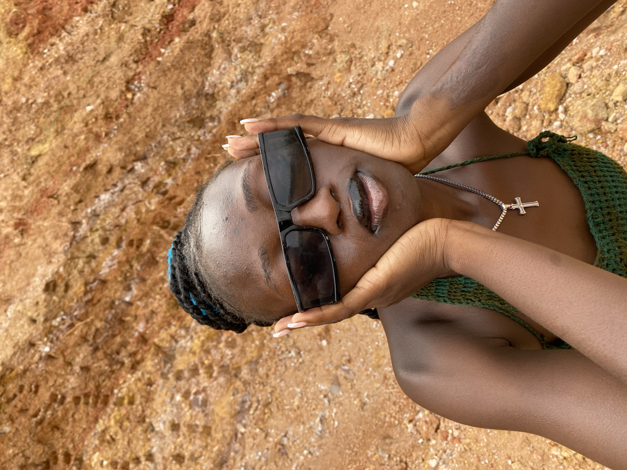 This media file features a close-up, top-down view of a young Black woman. She is lying on a sandy or rocky surface, with hands placed on either side of her head. She wears large, dark sunglasses and a green, crocheted bikini top. A silver chain with a cross pendant is visible around her neck. Her lips are slightly parted, and her eyes are closed or obscured by the sunglasses. The background is a textured, reddish-brown surface that appears to be natural earth or rock. The lighting suggests it is daytime, likely outdoors. The overall impression is one of relaxation or posing in a natural, possibly warm, environment.