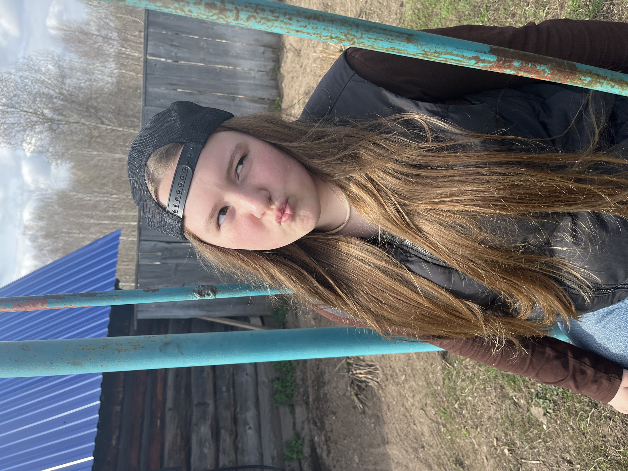 A young person, identified as female, is angled in the frame against a backdrop that suggests a rural or semi-rural setting. The person is wearing a black baseball cap, a black vest over a brown, long-sleeved shirt, and what appears to be a light blue garment underneath, possibly jeans. The individual is looking slightly upward and to the left, with pursed lips.
Behind the person, there is a wooden fence with what looks like a weathered appearance, suggesting an outdoor setting, which is probably the city of Буренка, Russia. There are also two rusted, blue metal bars, which may be part of a fence. A partially visible blue structure with angled planes is on the left. The ground appears to be dirt and sparse grass. Bare trees are visible in the distance.