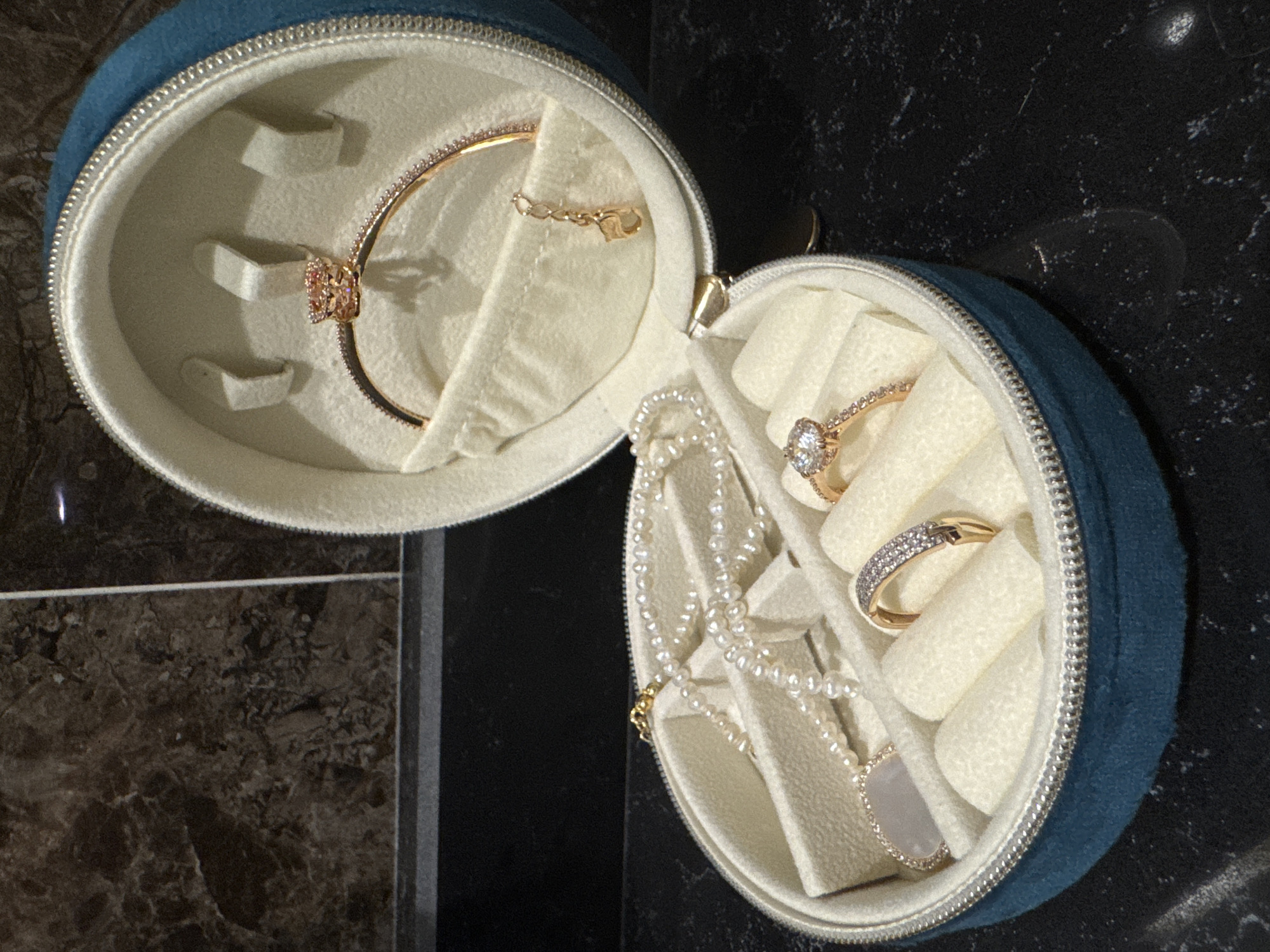 A round, blue jewelry case is opened to display its contents. The interior of the case is lined with a white, soft material with compartments for holding jewelry. The left side of the case holds a gold bracelet with a flower-shaped charm and a gold chain. The right side contains several rings, some with diamond accents, and a pearl necklace with a gold clasp. The case rests on a surface with a dark, marbled pattern. The scene is indoors.