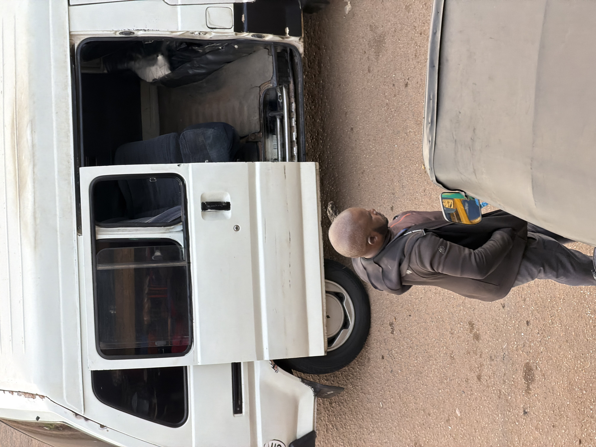 A man, bald and wearing a dark grey hooded sweatshirt and pants, stands beside two vehicles. He is looking down and to the right. One vehicle is a white van with its side door open, revealing dark seats inside. The other vehicle is a light grey car, partially visible, with a side mirror reflecting colorful objects. The scene appears to be on a paved road, with a rough asphalt surface. The lighting suggests it is daytime. There is no visible text in the image. The overall impression is a candid, everyday moment, possibly involving the man interacting with or preparing one of the vehicles.