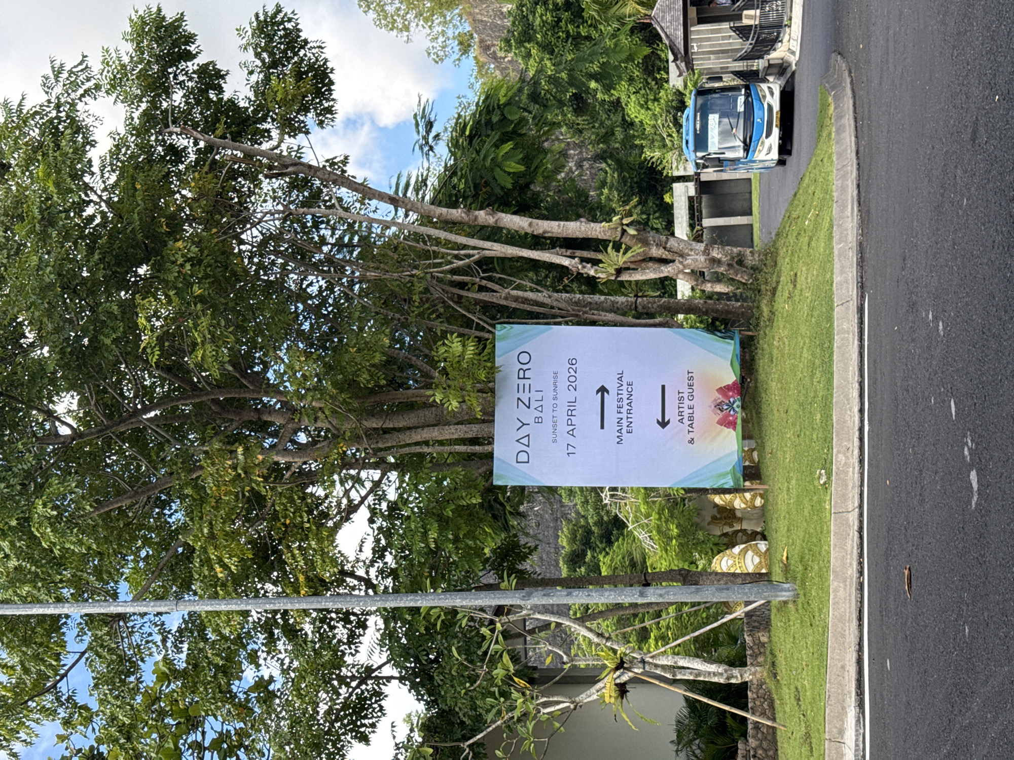 The image displays a bright, daytime scene in a lush, tropical environment, likely in Kuta Selatan, Indonesia. A prominent white sign, mounted on a metal pole, stands on a verdant grassy verge alongside an asphalt road.
The sign is a directional guide for an event, clearly stating "DAY ZERO BALI" and indicating the event takes place "SUNSET TO SUNRISE" on "17 APRIL 2026." It features two arrows: an upward arrow pointing towards the "MAIN FESTIVAL ENTRANCE" and a downward arrow for "ARTIST & TABLE GUEST." A colorful, abstract logo is present at the bottom right of the sign.
The setting is characterized by abundant green foliage. Numerous large trees with dense leaves and sturdy trunks occupy the left side and background, contributing to a vibrant, tropical atmosphere. The sky above is bright blue with scattered white clouds, indicating clear weather.
On the right side of the frame, a blue and white bus or shuttle vehicle is visible on the road, suggesting transport services, possibly for the event attendees or staff. In the background, hints of architectural structures with traditional-style roofs and stone steps can be seen, likely part of a resort or event venue. The scene depicts an entrance or access road for what appears to be a large-scale future event in a scenic location.