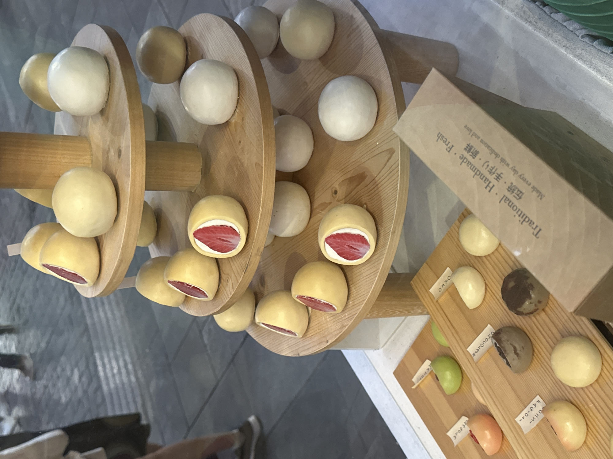The image captures a display of mochi in Milan, Italy. A wooden spiral structure holds mochi variations in white and yellow dough, some revealing strawberry filling inside. Nearby, a wooden display board presents different flavors like oreo, chocolate, matcha, and fragola, each marked with a label. A cardboard box labeled "Traditional · Handmade · Fresh" in both English and Japanese is also visible, indicating the fresh, artisanal nature of the mochi. The scene is situated indoors, likely in a shop, as evidenced by the reflective flooring and glimpses of clothing on people below the display.