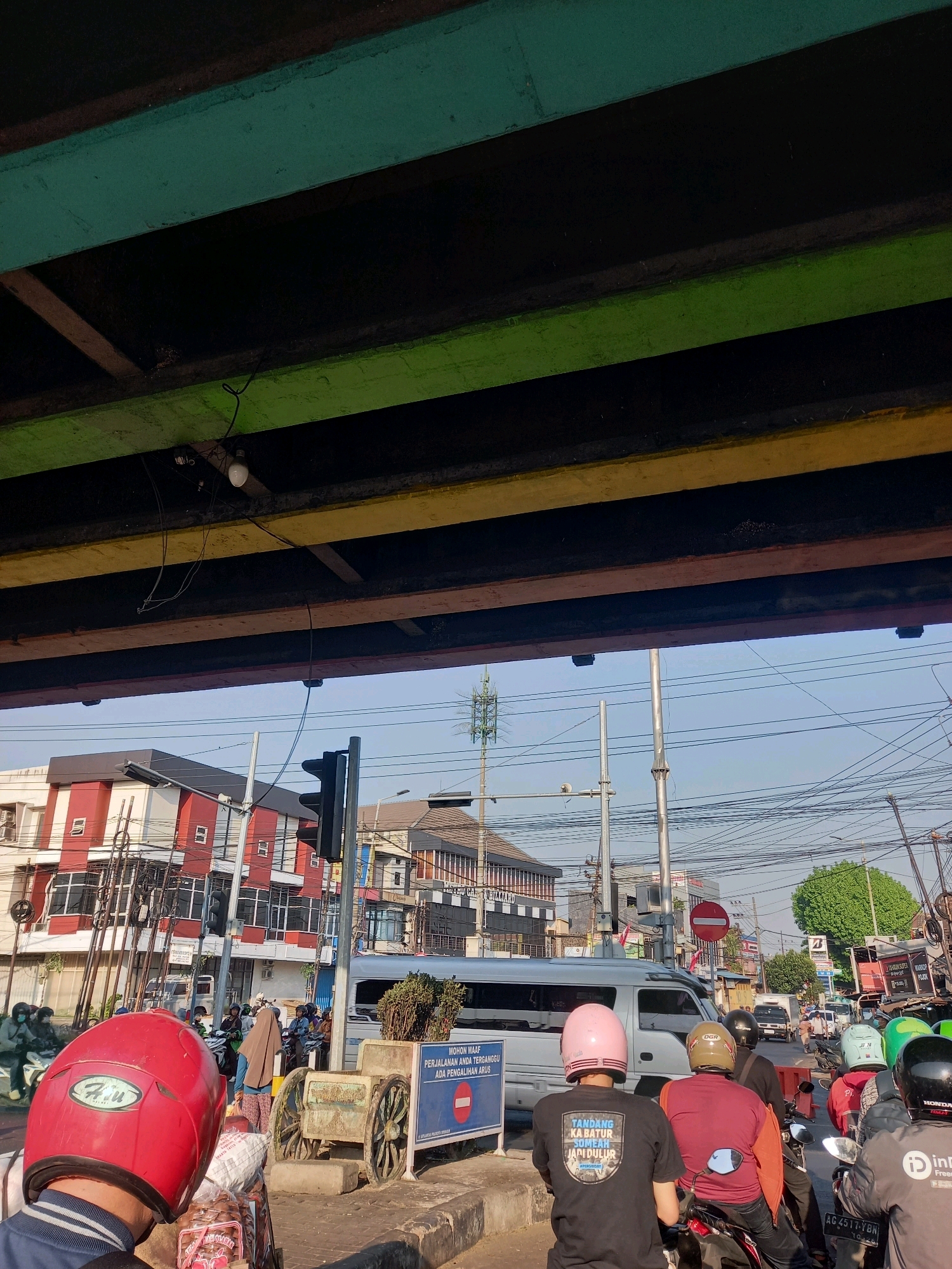 The image captures a street scene in Kedungrejo, Indonesia, from a low-angle perspective looking upwards towards a colorful overpass. The overpass consists of layered beams painted in green, black, and yellow, with a single light bulb hanging from the underside of one of the beams. Below the overpass, a busy street is visible.
Several individuals are on motorcycles, some wearing helmets. In the foreground, the back of a person wearing a red helmet is prominent. To the left, buildings with red and white striped facades line the street, with multiple power lines crisscrossing the sky above. A traffic light stands at an intersection, currently showing a black signal head. A white van is stopped in traffic, and a signpost with Indonesian text and a red circular "no entry" symbol is visible. In the middle ground, a cart with large wooden wheels is parked near a sign. Further down the street, more vehicles and people are present, and a tall communication tower can be seen in the distance. The overall atmosphere suggests a sunny day in an urban environment with significant traffic.