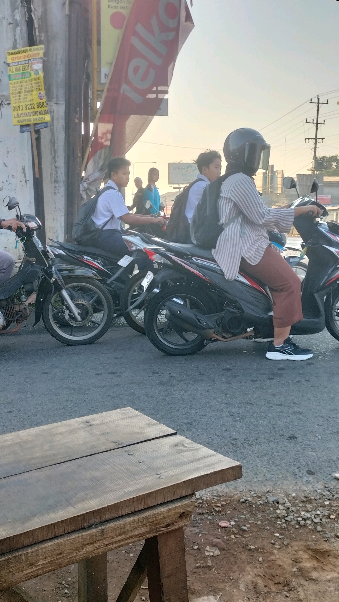 The image depicts a street scene in Subah, Indonesia, with several people on motorcycles. In the foreground, a rustic wooden table is positioned on a dirt and gravel area next to a paved road. Behind the table, multiple motorcycles are stopped or moving slowly, creating a traffic scene.
On the motorcycles, there are at least four individuals. Two young males wearing white shirts and dark backpacks are seated on separate motorcycles. One of them appears to be looking forward, while the other is also looking forward. To their right, a woman wearing a striped long-sleeved shirt and brown pants is riding a motorcycle. She has a black helmet with a transparent visor, obscuring her face. She is looking forward, and her right hand is on the handlebar. Another person, possibly male, is partially visible behind her, also on a motorcycle. The motorcycles themselves are modern scooters.
The environment includes a roadside with a tall pole supporting a large banner that appears to be an advertisement for "Telkomsel." Another smaller yellow flyer with text and a phone number is attached to a pole on the left side of the frame. In the background, there are buildings and utility poles, indicating an urban or semi-urban setting. The sky is visible and appears to be during daylight, possibly late afternoon or early morning, with soft lighting. The overall scene suggests a typical street environment in Indonesia with common modes of transportation.