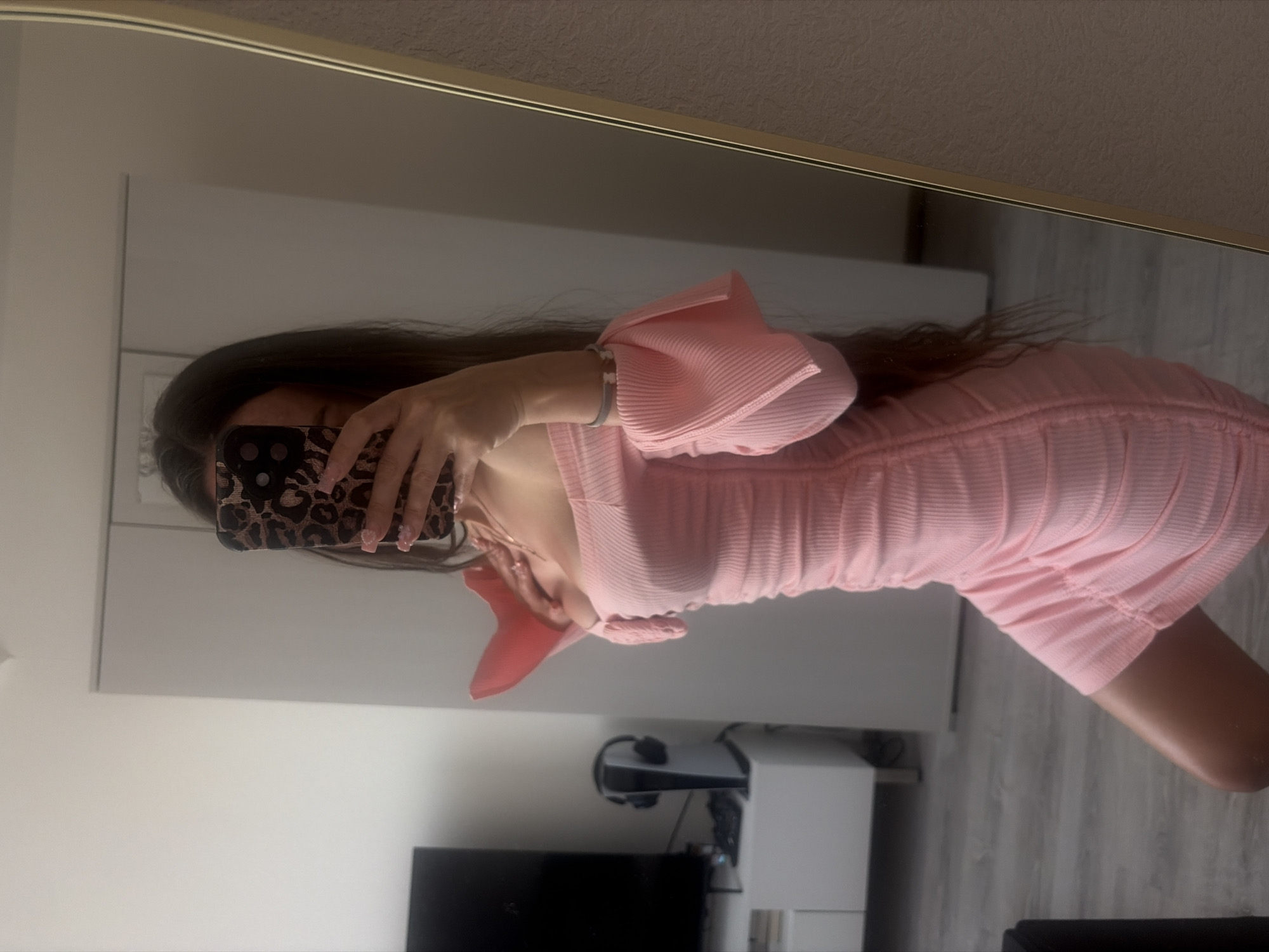 A person stands in a room, taking a mirror selfie. They are wearing a pink, off-the-shoulder, ruched dress with bell sleeves. The person's phone, with a leopard-print case, is held up to cover their face. The room appears to be a bedroom or dressing area, with a white wardrobe and a dresser with a television and gaming console visible in the background. A tall, gold-framed mirror dominates the right side of the image. The floor is wood-effect laminate. The overall lighting suggests it is daytime.