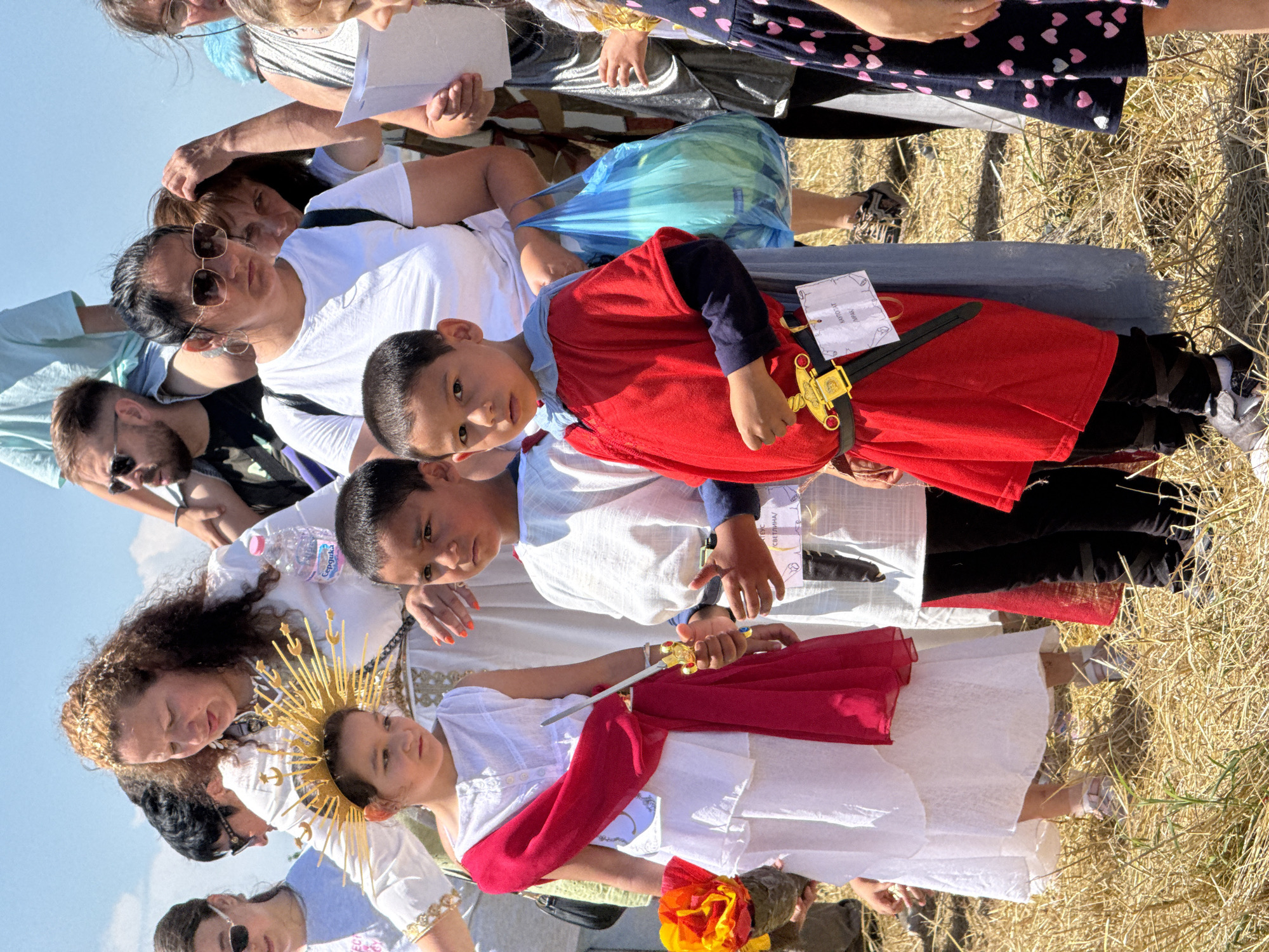 The image depicts an outdoor scene in Gigen, Bulgaria, featuring a gathering of individuals, predominantly adults and children, in a dry, grassy field under bright daylight. In the immediate foreground, a young girl, approximately 6-8 years old, stands on the left. She wears a layered white dress, a bright red sash draped from her left shoulder across her torso, and a prominent golden sunburst-style headpiece. She holds a small, ornate silver-colored sword with a gold hilt in her right hand, pointed downwards. In her left hand, she holds a prop resembling a torch with orange and yellow fabric "flames." Her expression is focused, looking slightly down and to her right. To her right stands a young boy, also approximately 6-8 years old. He is dressed in a red tunic over a dark blue long-sleeved shirt, with a light blue fabric draped over his shoulders. A black belt with a prominent gold, ornate buckle cinches his waist, from which a black sheath containing a small sword with a gold hilt is suspended. A white paper tag with Cyrillic text, including "КАРПОТАТ" and "ДРАЖАР," is attached to his belt. He looks directly forward with a serious expression. Behind the two children, a woman with curly, light brown hair wears a long white dress with gold embroidered trim on the sleeves and hem, and a simple gold necklace. A smaller golden headpiece is visible in her hair. She holds a plastic bottle of "Сердика" brand water in her left hand and looks down towards the children. Her fingernails are painted bright orange. Further back and to the right of the central woman, another woman with dark hair and dark sunglasses wears a white t-shirt and carries a dark backpack. She holds a blue plastic bag containing a light green object. Partially obscured behind her, another woman with reddish-brown hair is visible, touching her head. On the far right, an older woman with glasses is seen holding white papers. In the background, other adults are visible, some partially obscured or out of focus. On the far left, a woman with sunglasses and a light blue t-shirt is partially visible. On the upper right, a man with sunglasses, wearing a dark t-shirt, appears to be carrying a child on his shoulders, though only the child's lower legs are visible. The ground is covered with dry, light brown straw or dead grass, suggesting a summer or