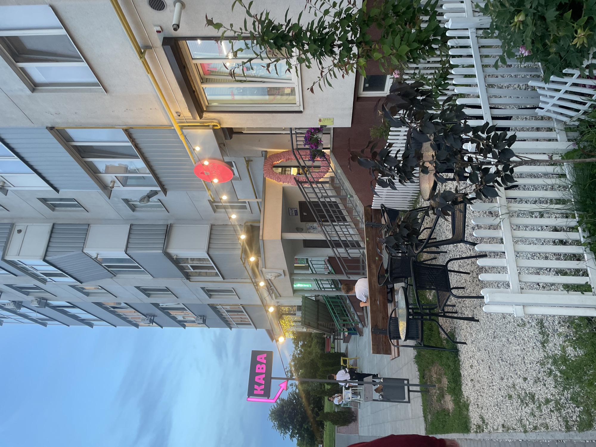 The image depicts an outdoor cafe area and the entrance to a multi-story building in Rozsoshentsi, Ukraine, likely during dusk or early evening. The scene is illuminated by string lights and neon signs.
On the left, a modern multi-story building with multiple windows and balconies dominates, featuring light grey and darker paneled sections. A yellow pipe runs along its exterior. The cafe occupies the ground floor of an adjacent building on the right, which has a brown facade.
The cafe's outdoor seating area is visible in the foreground and to the right. It features several metal tables and chairs set on a gravel surface. A white picket fence separates this area, and green bushes and plants are scattered around. A prominent pink neon sign in the foreground reads "KABA" (likely "Кава," meaning "Coffee" in Ukrainian) with an arrow pointing towards the cafe entrance. Another circular pink sign with text, possibly "АКЦІЯ" (promotion), hangs above the entrance.
A decorative pink, heart-shaped garland adorns the railing of the stairs leading up to the cafe entrance. String lights are strung across the facade, casting a warm glow. Several people are present: one person in a white shirt is seated at a wooden table near the cafe entrance, and two other individuals are seated further to the left in the outdoor area, near the "KABA" sign. They appear to be relaxing or socializing. The sky above is a clear, soft blue, indicative of twilight.