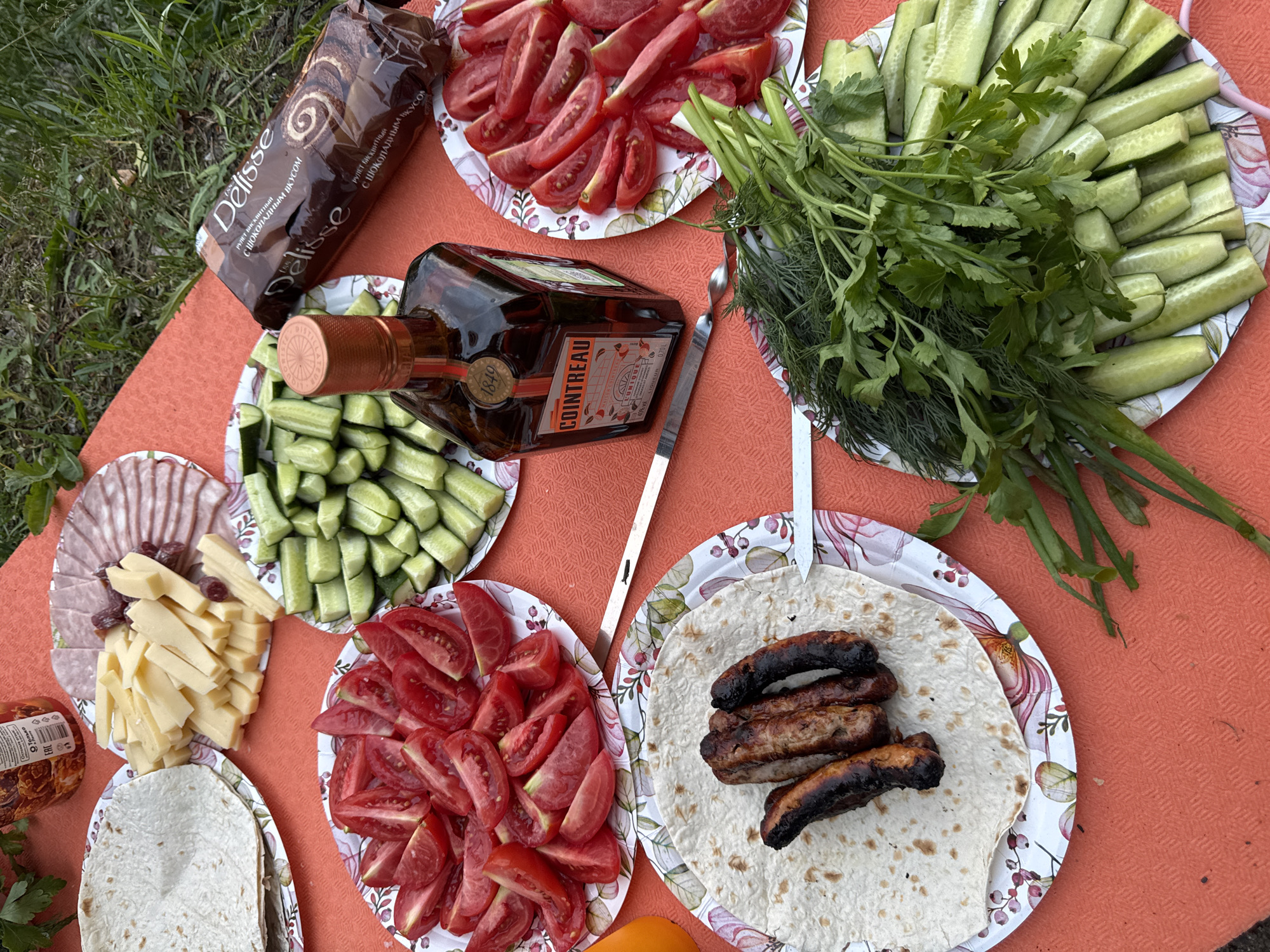 The image depicts an outdoor picnic laid out on an orange tablecloth, likely in the city of Bolshaya Izhora, Russia. Several plates are filled with food, including sliced tomatoes, cucumbers, cheese, and a plate of sausages on tortillas. There is also a bottle of Cointreau liqueur and a package of Delisse chocolate roll. A skewer is visible among the food items. The setting appears to be a casual gathering, possibly a barbecue or a picnic on a grassy area. The overall atmosphere is relaxed and suggests a meal being prepared or enjoyed outdoors.