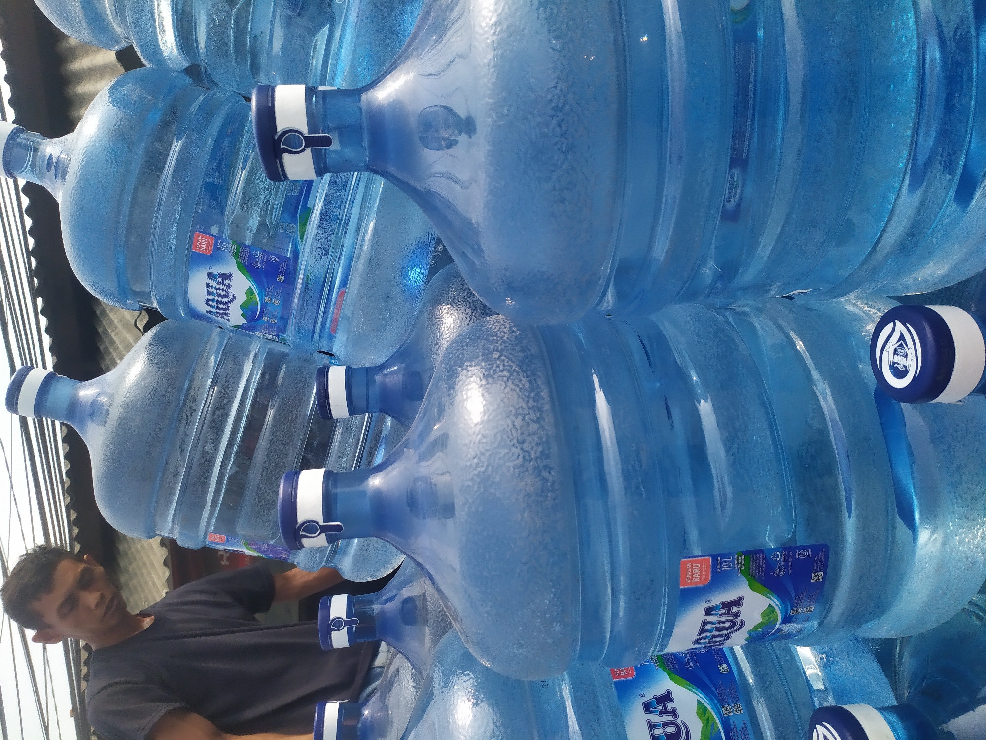 In the city of Kragilan, Indonesia, a man in a dark t-shirt stands behind a large stack of Aqua brand 19-liter water bottles. The bottles, made of clear blue plastic, are arranged in several rows, some partially obscured by others. The man is looking down and to his left, with his face partially in shadow. The background consists of a corrugated metal roof and overhead wires, suggesting an outdoor or semi-outdoor setting. The lighting is bright, casting subtle reflections on the curved surfaces of the water bottles. The overall impression is of a delivery or storage area for bottled water.