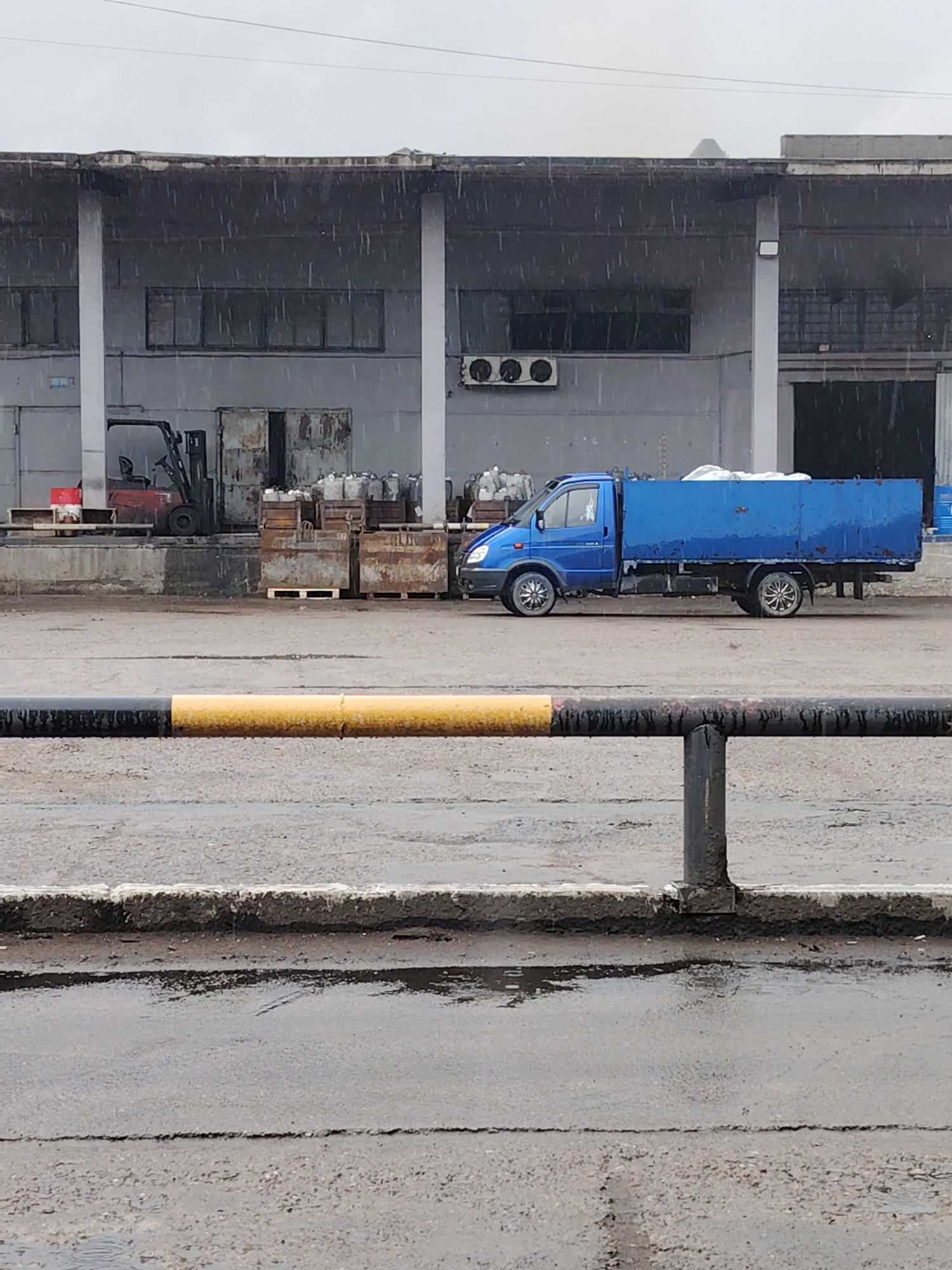 This image captures an industrial or commercial loading area in Noviy, Russia, on a rainy and overcast day. Heavy rain is visibly falling, creating streaks in the air and leaving the asphalt ground wet with numerous puddles.
In the midground, a bright blue flatbed truck, likely a GAZelle, is parked on the damp pavement. It features distinct chrome hubcaps and appears to have a tarp or some items covered in white in its bed. To the left of the truck, positioned near a loading bay of the building, stands a red forklift. The area around the loading dock is cluttered with various rusty metal containers, wooden pallets, and white storage bins.
The background is dominated by a large, grey industrial building or warehouse. It has a practical, utilitarian design with multiple loading bay doors, some small, dark windows, and several air conditioning units mounted on its facade. The roofline shows signs of age and wear.
In the foreground, a black and yellow painted metal barrier or railing spans horizontally across the lower part of the frame, separating the viewer from the industrial scene. The ground in front of the barrier is rough, cracked asphalt. No people are visible, and no discernible text can be read on any of the objects or structures. The scene conveys a functional, somewhat dreary industrial environment under wet weather conditions.