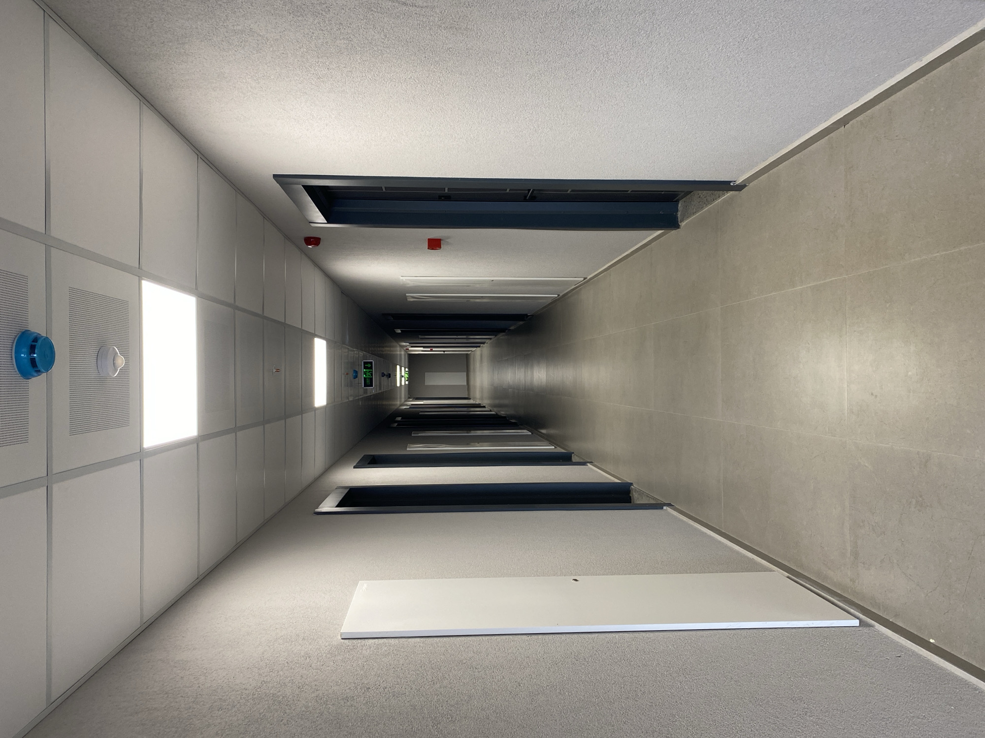 The image depicts a long, empty, interior hallway with a tiled floor and textured gray walls. The hallway is lit by fluorescent ceiling panels. There are several doors visible on both sides, and green exit signs with illuminated figures are mounted on the ceiling towards the end of the hallway. A blue smoke detector is visible on the ceiling near the top of the image. The scene appears to be a corridor in a modern building, possibly a hotel, office building, or residential complex, located in Batumi, Georgia, given the context provided. There are no people or visible actions taking place in the hallway.