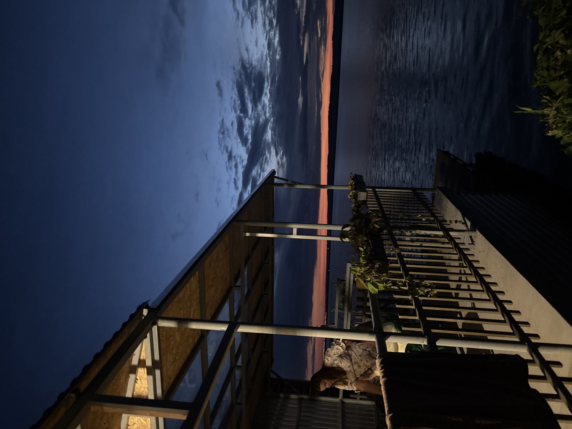 The image depicts a waterfront view at dusk or dawn from a covered balcony or deck. A woman in a floral-patterned dress is standing on the balcony, looking down. The balcony has a metal railing with some plants in planters and a dark-colored towel or blanket hanging on the railing. The balcony structure has a metal frame and a light-colored roof. Beyond the balcony, a large body of water, likely a river or lake, stretches to the horizon. The sky is a gradient of deep blue and orange, with scattered clouds, indicating twilight. In the distance, the silhouette of a distant shoreline or city lights is visible. This scene is consistent with the waterfront area of Chaikovsky, Russia, known for its location on the Kama River reservoir.