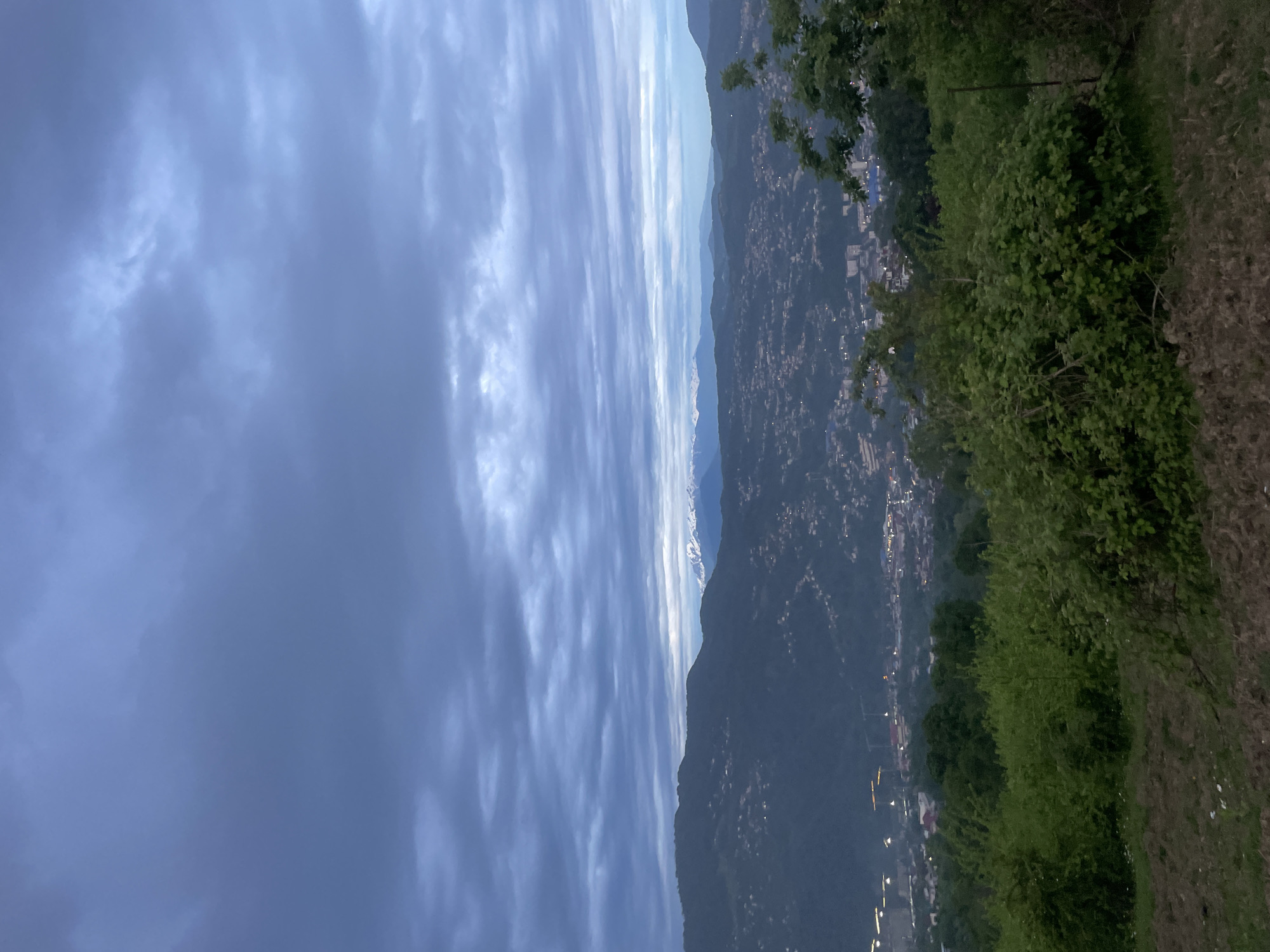 The image is a landscape view taken from a high vantage point, possibly a hilltop or mountain overlooking a valley or town. The sky is dominant, displaying streaky clouds that create a linear pattern across the frame. Below the sky are mountains, and a cityscape is nestled within the mountains. The terrain is lush with greenery, and a shrubbery appears in the foreground. The image suggests a location with significant elevation changes and a mix of natural landscapes and urban development. Baranovka, Russia is not visible in the image.