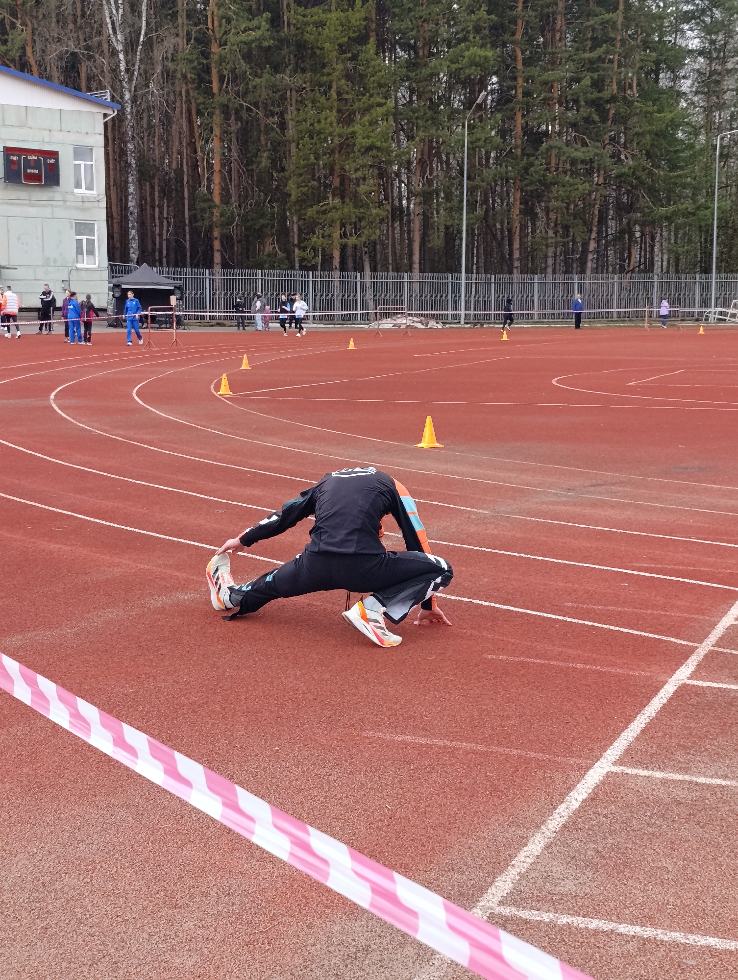 The image shows a track and field stadium in the city of Chaikovsky, Russia. A person in black athletic wear with orange accents is stretching on the red running track, near a pink and white striped barrier tape. Other people are visible in the background of the track near the track house. Several orange cones are set up around the track. Tall pine trees are in the background, along with a building with a scoreboard.