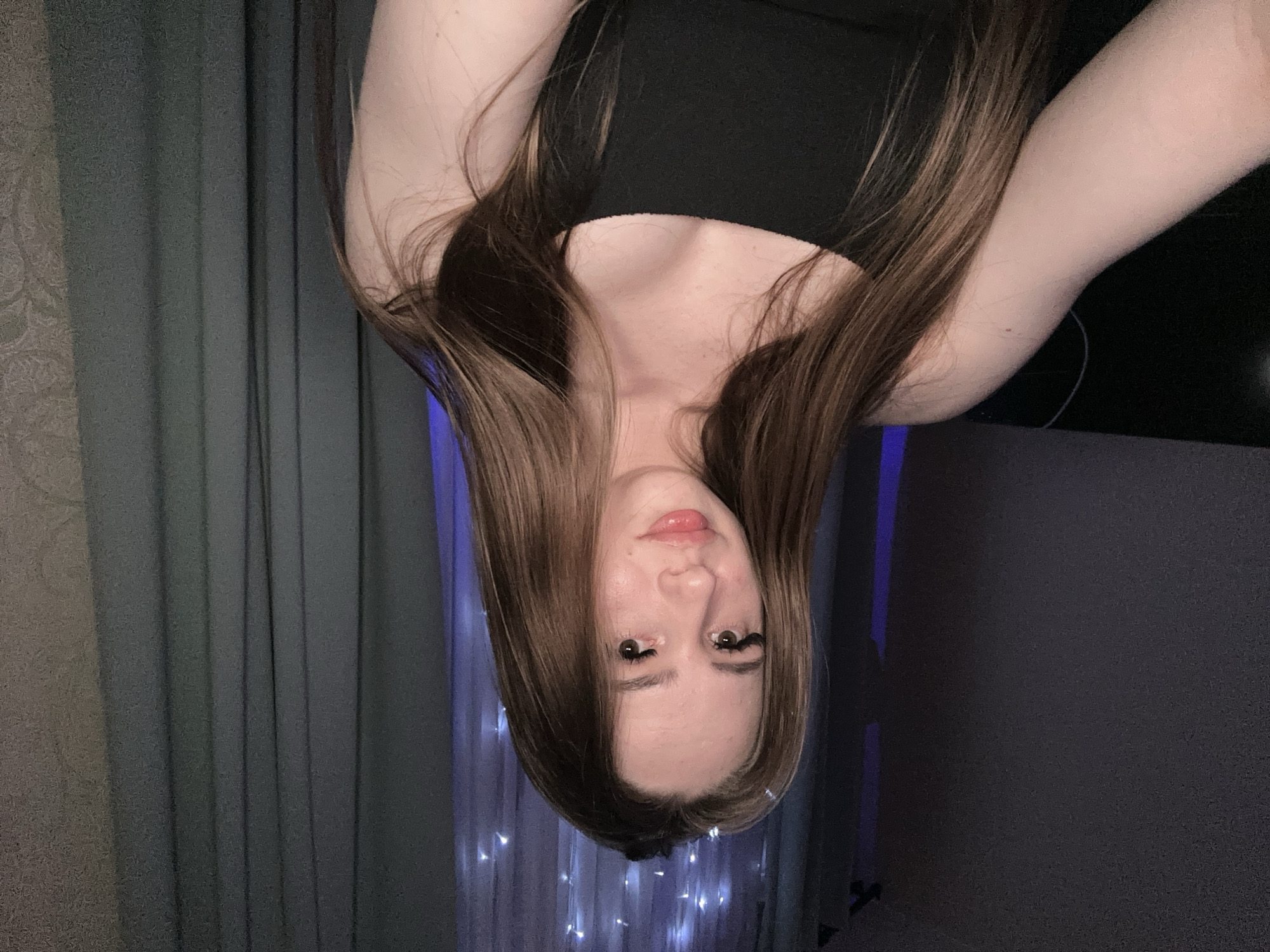 A photo depicts a person with long brown hair wearing a black top. The person is oriented upside down in the frame. The background features curtains and a dark surface. The location is not specified.