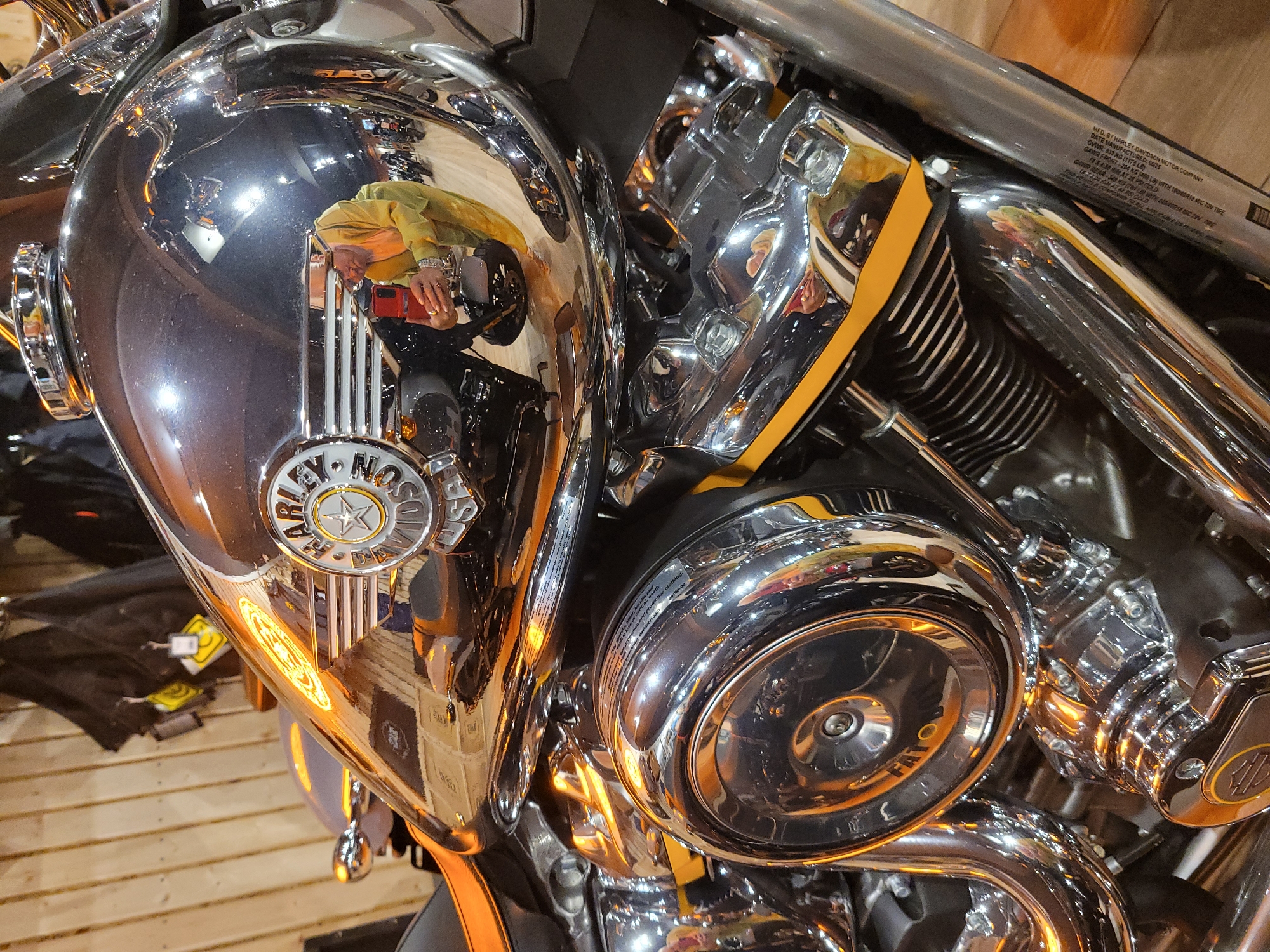 A close-up shot focuses on the reflective chrome gas tank and engine components of a Harley-Davidson motorcycle. The Harley-Davidson logo is prominently displayed on the gas tank. Reflections on the chrome surfaces reveal a person taking a picture of the motorcycle with a phone. A wooden wall is visible in the background. The scene depicts details of the motorcycle and the surrounding environment. There is no indication of Moscow, Russia.