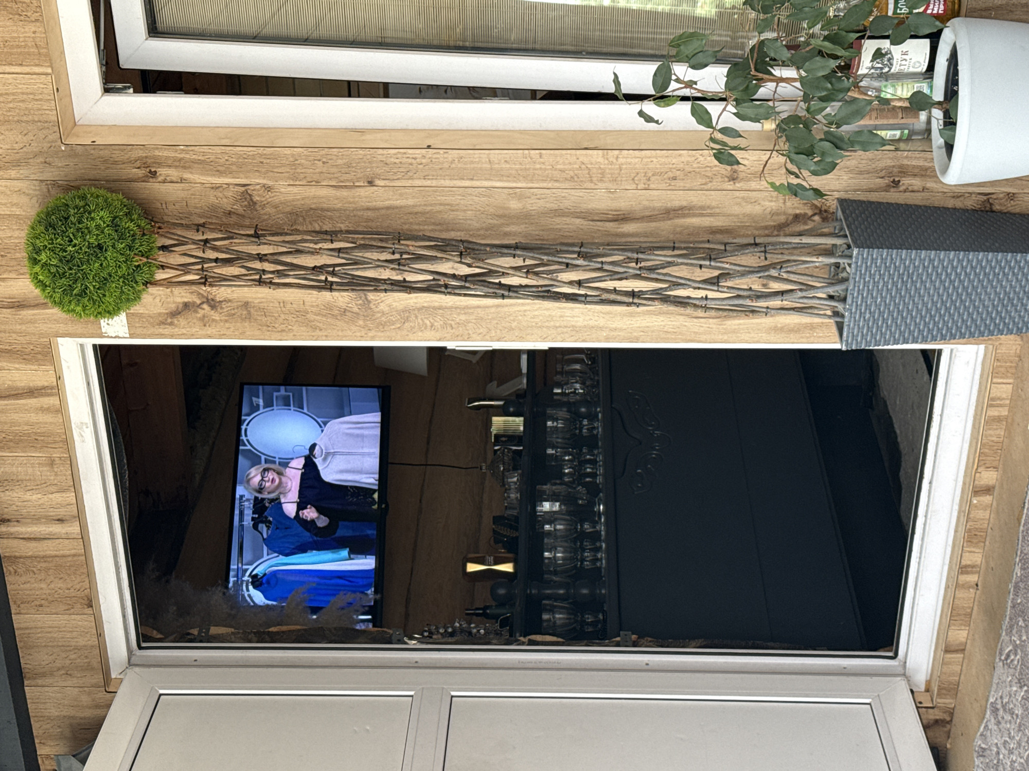 The image depicts a covered outdoor space with wooden plank walls and a white-framed door. Inside the doorway, a television displays a woman with glasses wearing a black off-the-shoulder top and holding a grey cardigan. Various garments, including blue tops, are hanging on a rack behind her. Below the television, a dark cabinet is visible, filled with glassware and decorative items. To the right of the door, a tall, grey, woven planter holds a decorative topiary sphere. Further to the right, a smaller potted plant with green leaves sits next to several bottles, including one with a label that appears to be in Cyrillic. The ground in front of the door is covered with a textured mat. The overall impression is of a residential outdoor area, possibly a porch or patio, with elements of home entertainment and decoration.