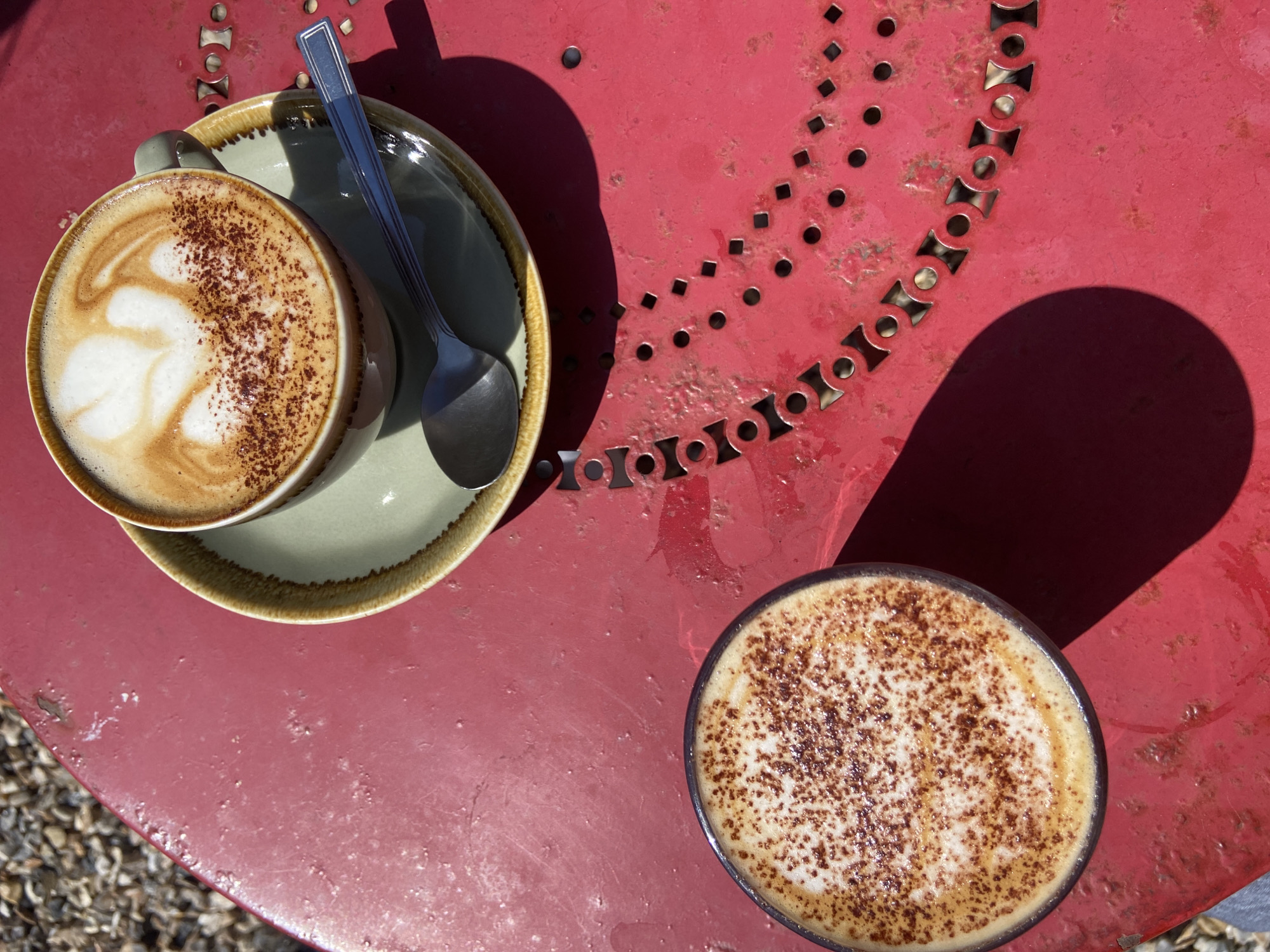 Two cups of cappuccino sit on a red metal table in London, United Kingdom. One cappuccino rests on a light green saucer with a silver spoon. The cappuccino shows latte art. The table has a decorative pattern of cutouts. Gravel is visible at the edge of the frame. The light suggests it is daytime.