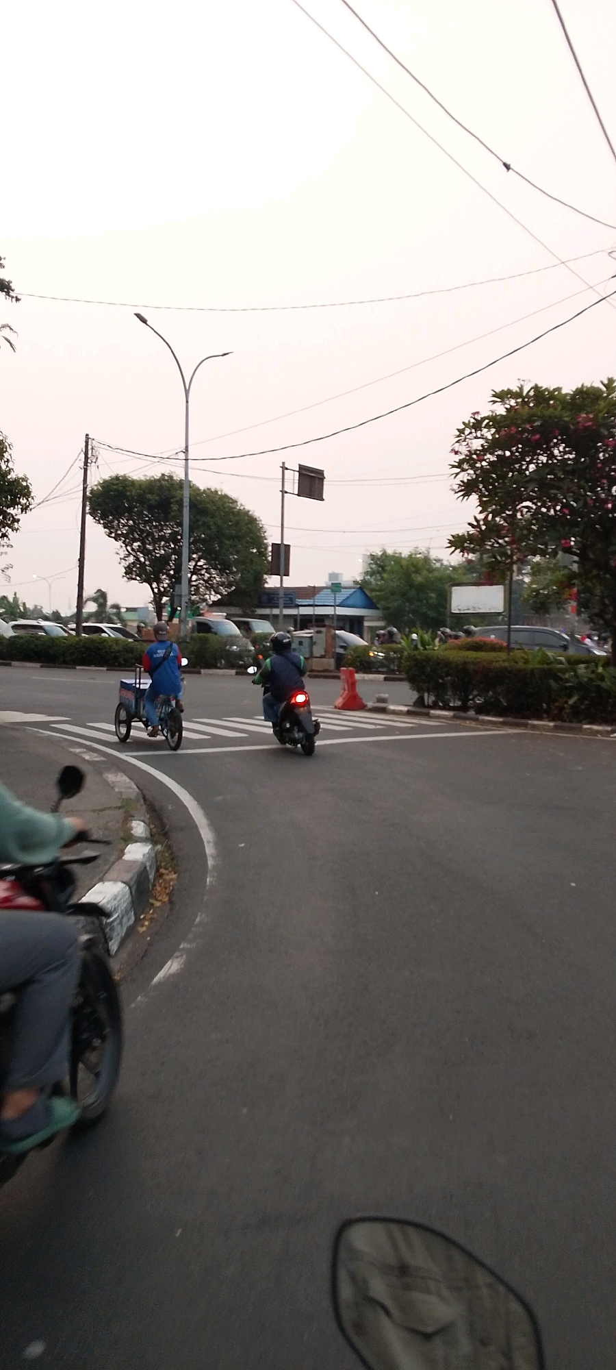 The image depicts a street scene in Depok, Indonesia, likely during the day under an overcast sky. In the foreground, a person is riding a motorcycle, with their legs and the motorcycle's handlebars visible. Further down the road, another motorcycle is seen from behind, ridden by an individual wearing a helmet and a dark vest over a lighter shirt, with a backpack. This motorcycle is positioned on a crosswalk. To the left of the crosswalk, a person is riding a bicycle-powered cart, also wearing a helmet and a blue shirt with a backpack. The cart appears to be a mobile food or goods vendor, with a white box on its rear.
The road surface is asphalt, with white pedestrian crossing lines and a white curb marked with black and white stripes. On the right side of the road, there are orange traffic cones and dense green bushes. Beyond the road, there are parked cars, trees with green foliage, and some buildings in the distance. Utility poles and overhead wires crisscross the sky, which is a light, diffused color, suggesting a cloudy or hazy atmosphere. A street lamp is visible on the left side of the frame. The overall environment appears to be an urban or suburban area with moderate traffic.