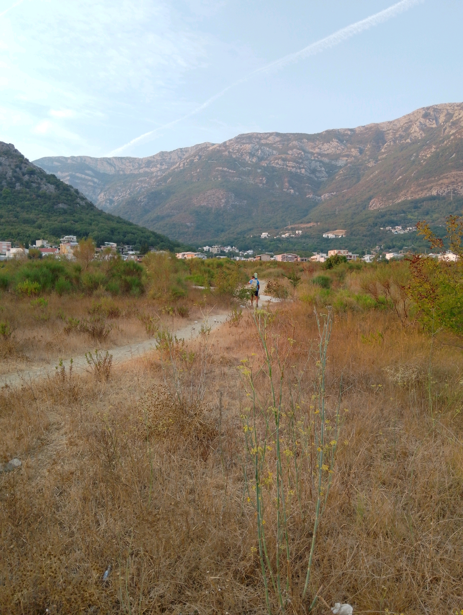 A person wearing a blue and white shirt and dark shorts walks on a dirt path through a dry, grassy field. In the background, a town with white and pink buildings is nestled at the base of a lush green mountain range. The sky above is a pale blue with a thin white contrail. The image is taken during the day, likely in the late afternoon given the soft lighting. The scene appears to be in the Sutomore area of Montenegro.