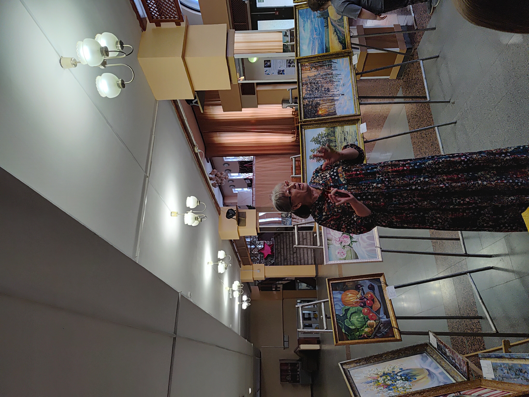 The image shows an indoor art exhibition. A woman in a floral dress is standing and gesturing as if speaking to an audience. Several framed paintings are displayed on easels in front of her, depicting still lifes, landscapes, and floral arrangements. In the background, there are stairs, curtains, and lighting fixtures on the ceiling. The flooring appears to be polished concrete or tile. The overall environment suggests a community center or a small gallery space. The specific location in Марковский, Russia is not identifiable from the visual cues alone.