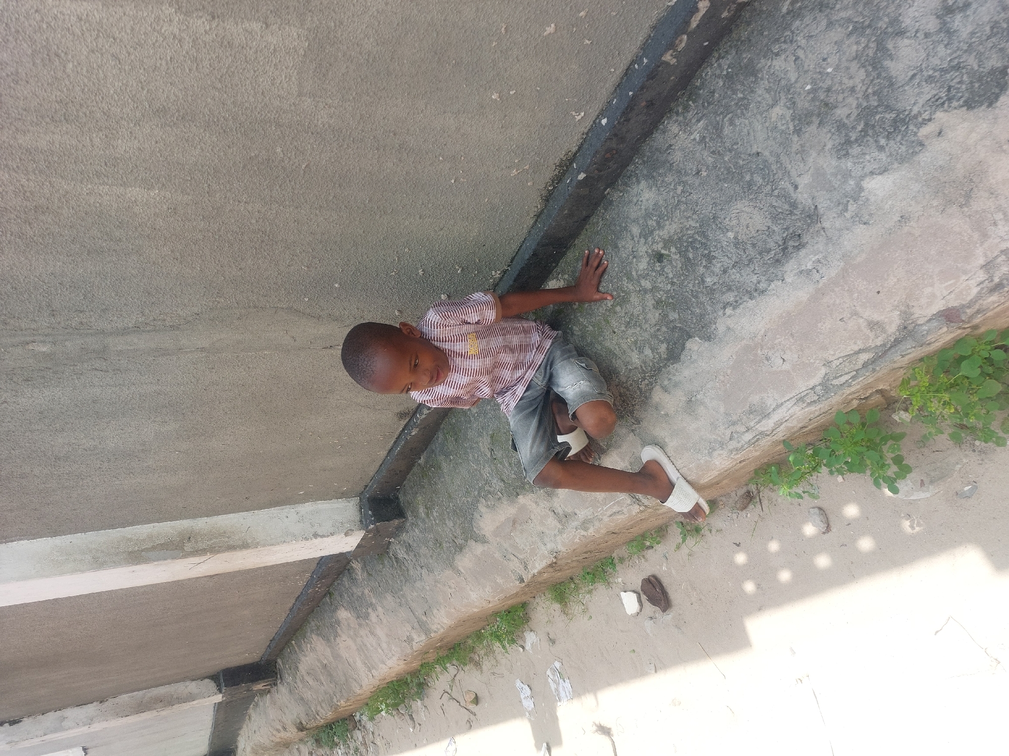 A young boy in a striped shirt, denim shorts, and white sandals is on a concrete structure that appears to be the exterior of a building. He has one hand on the concrete. There is sand and some vegetation adjacent to the structure. This is likely Dar es Salaam, Tanzania.