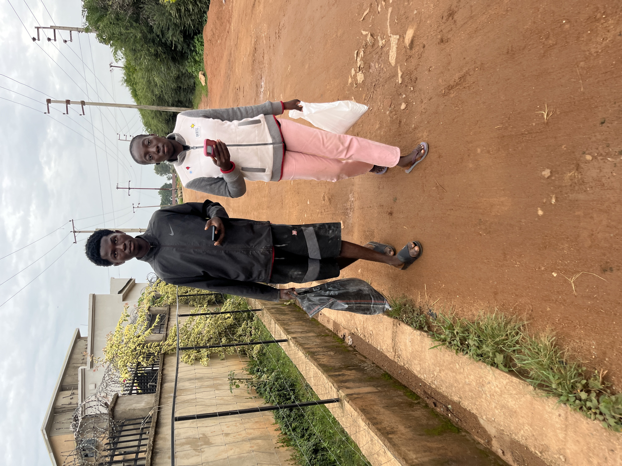 Two individuals, a young Black male and a young Black female, walk side-by-side on a dirt road in Bukuru, Nigeria. The male, on the left, wears a black jacket with a Nike logo, black shorts with grey trim, and black slide sandals. He holds a black plastic bag in his left hand and a smartphone in his right. The female, on the right, is dressed in a light pink vest with grey sleeves, a red collar trim, and light pink pants. She wears dark slide sandals and holds a pink smartphone in her right hand and a white plastic bag in her left. The road is unpaved and appears well-traveled. To the left of the road, a concrete drainage channel is visible, bordered by sparse green vegetation and a low concrete wall. Beyond the wall, a building with a fenced perimeter topped with barbed wire is seen, along with some flowering bushes. Power lines crisscross the overcast sky. The environment appears somewhat arid, with dry earth and limited greenery.