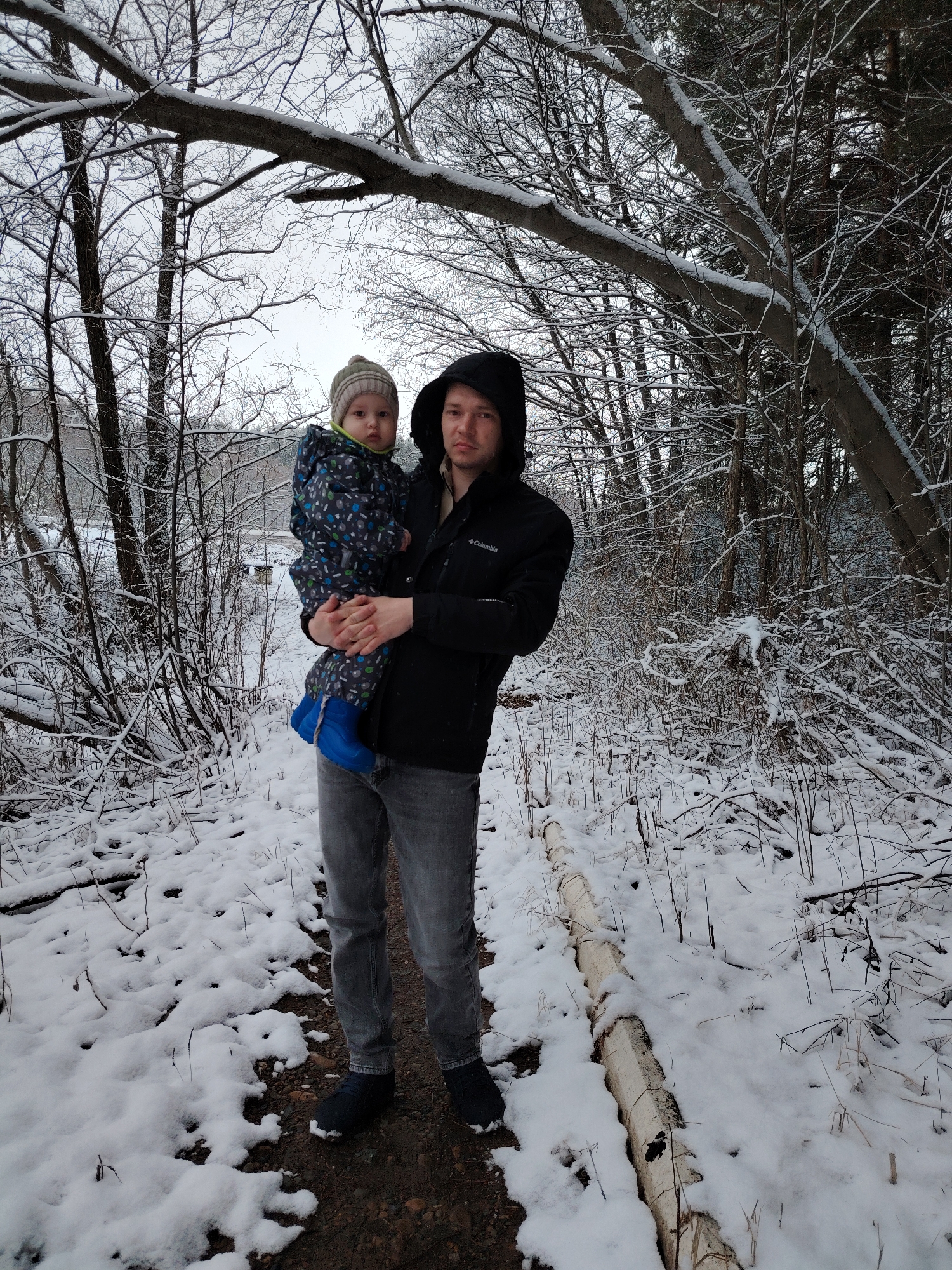 The image captures a man holding a young child in a snowy forest setting, potentially located near the city of Большая Уса, Russia. The man is wearing a black hooded jacket and gray jeans. The child is clad in a patterned snowsuit, a knitted hat, and blue boots. Both are facing the camera. The background features snow-covered trees and branches, suggesting a winter season. A fallen birch log covered with snow is on the ground next to the man. The scene lacks explicit urban elements, suggesting an immediate natural environment.